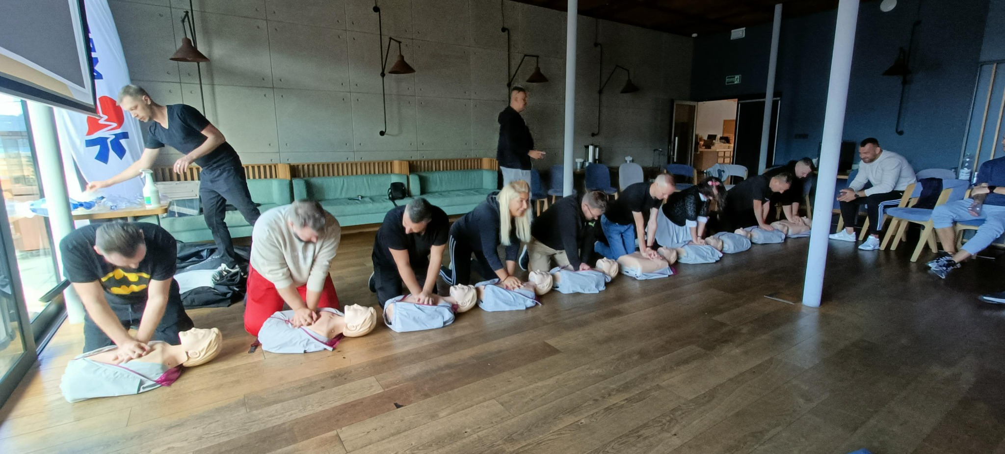 Uczestnicy szkolenia w Łodzi ćwiczą resuscytację krążeniowo-oddechową na manekinach na tle betonowej ściany i drewnianej podłogi.