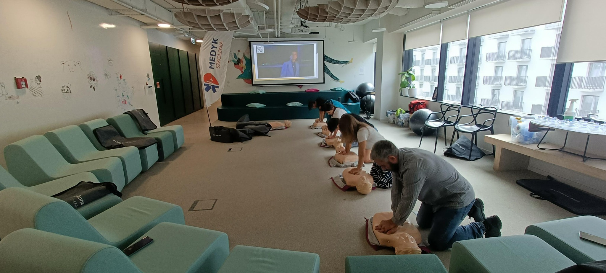 Uczestnicy kursu pierwszej pomocy ćwiczą resuscytację krążeniowo-oddechową na manekinach w przestronnej sali szkoleniowej z projektorem i flagą Medyk Szkolenia.