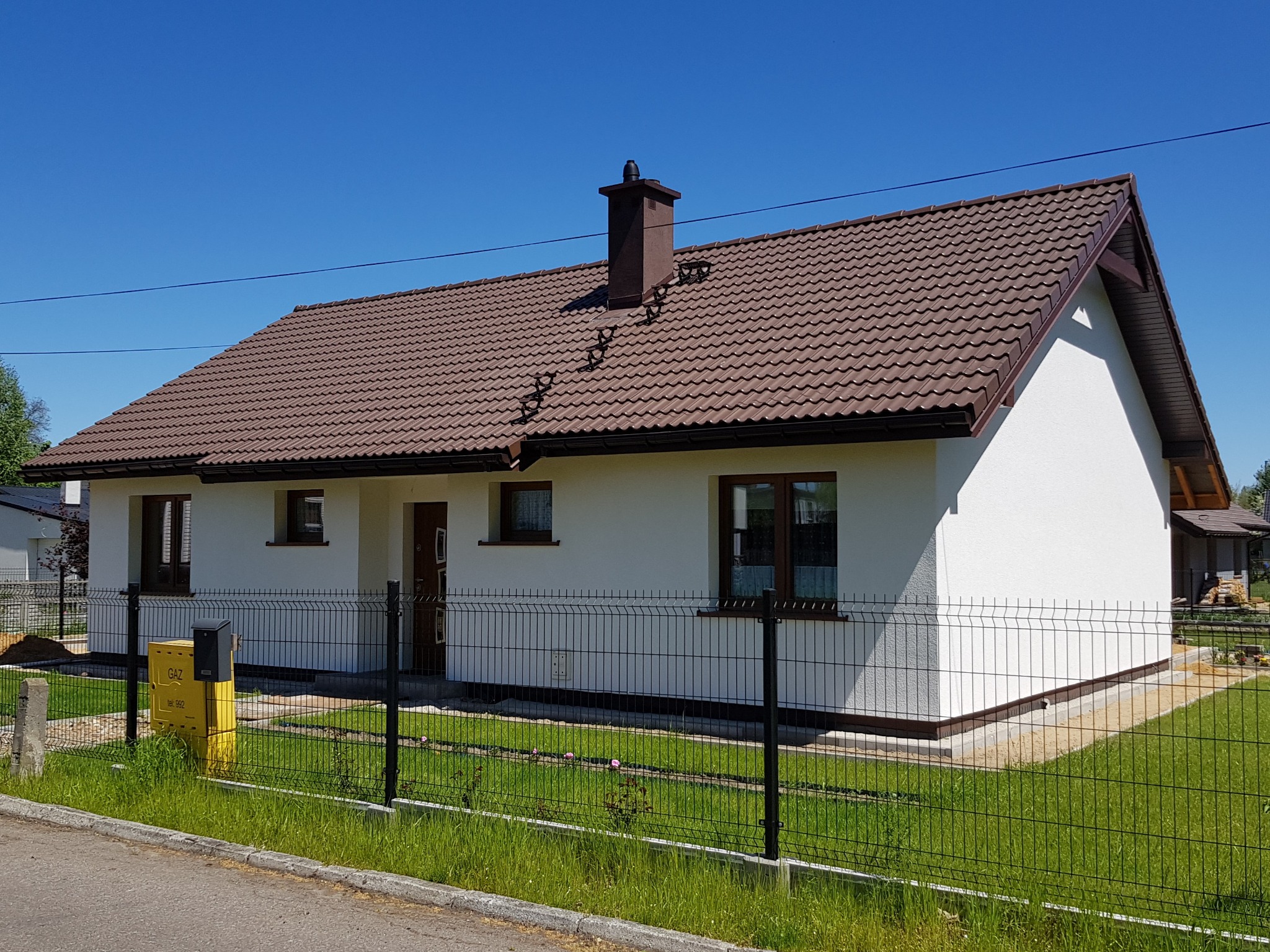 Parterowy dom jednorodzinny z brązowym dachem i kominem, ogrodzony czarnym metalowym płotem z żółtą skrzynką gazową na pierwszym planie.