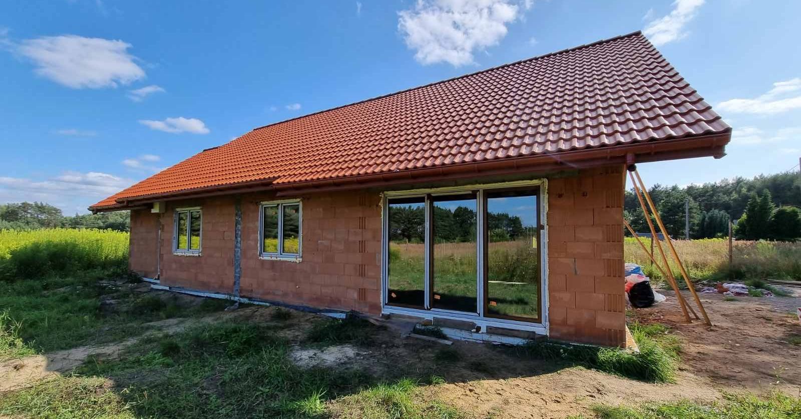 Niedokończony dom murowany z czerwoną dachówką, widoczne okna z PCV, ściany z pustaków ceramicznych, teren wokół domu zarośnięty trawą.