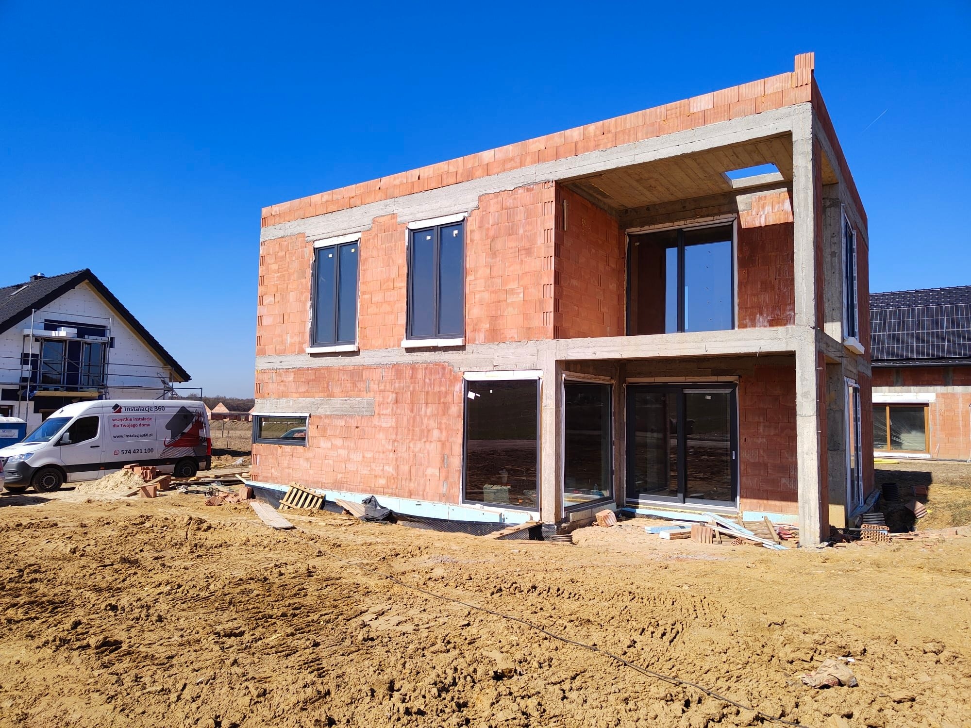 Surowy stan dwukondygnacyjnego domu z czerwonej cegły z zamontowanymi oknami, na pierwszym planie nierówny teren budowy i furgonetka firmy instalacyjnej w tle.