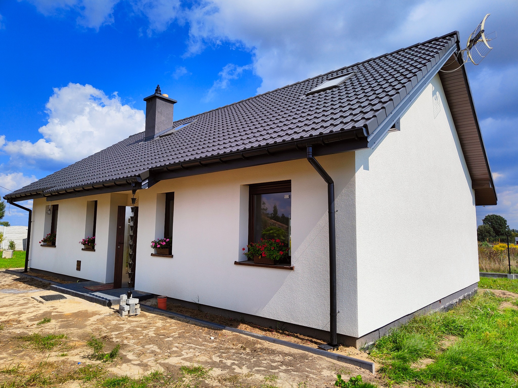 Niedawno ukończony, parterowy dom z białymi ścianami, ciemną dachówką i oknami z kwiatami, widoczne ułożone bloczki betonowe przed wejściem.