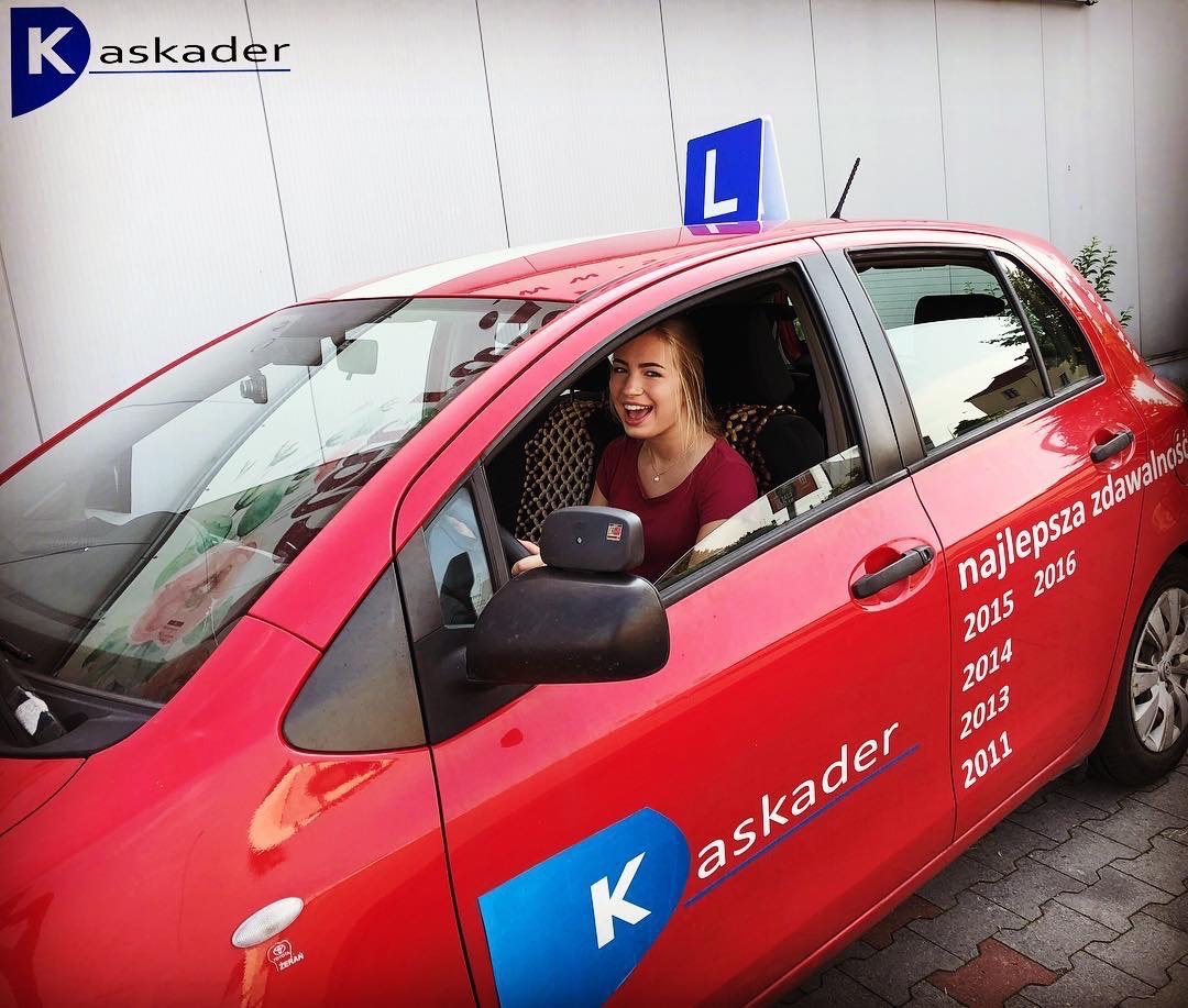 Uśmiechnięta młoda kobieta siedzi za kierownicą czerwonego samochodu z niebieską literą 'L' na dachu, oznaczającego naukę jazdy. Na drzwiach widnieje napis 'najlepsza zdawalność' z latami 2011-2016...