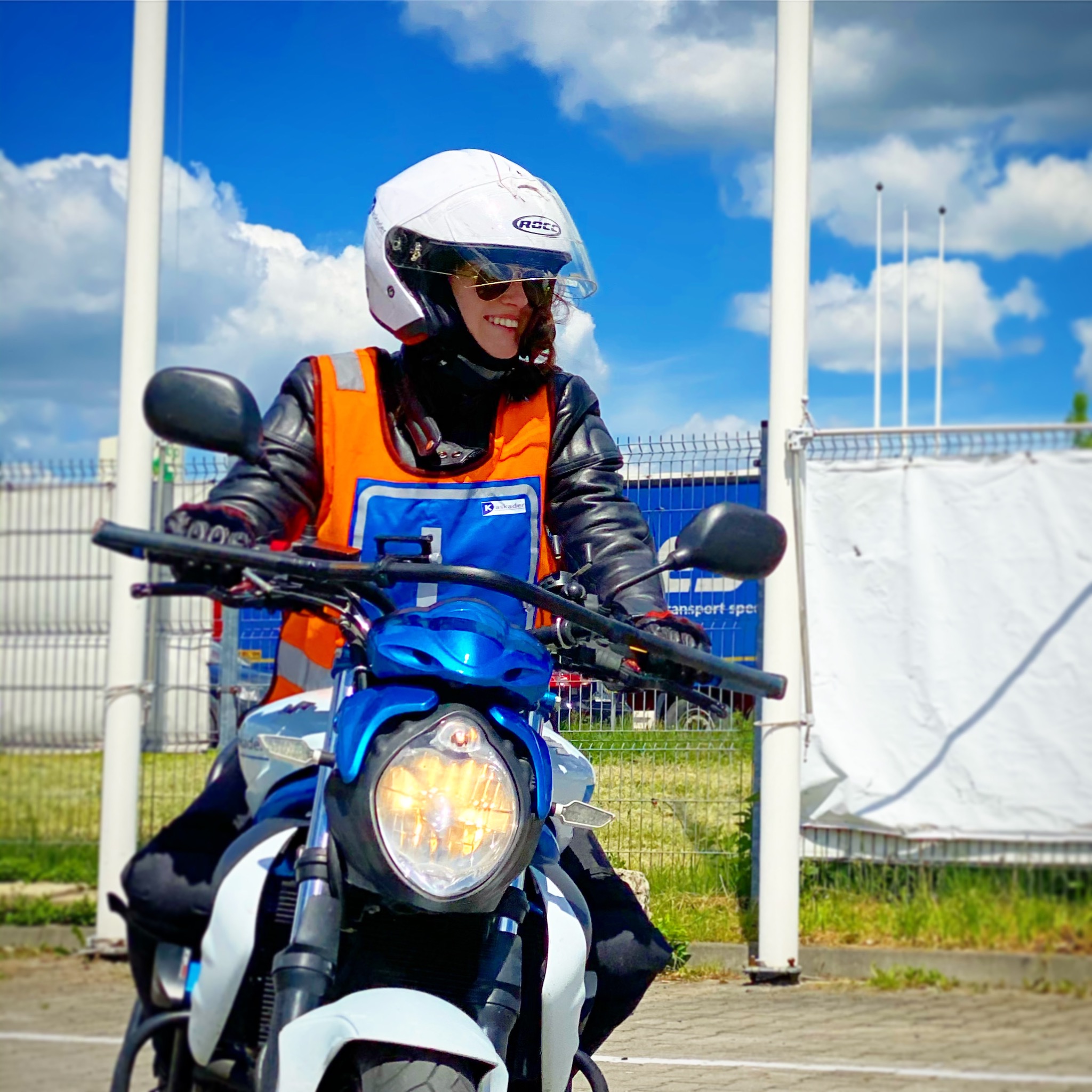 Uśmiechnięta kobieta w kasku i kamizelce odblaskowej siedzi na motocyklu, na tle błękitnego nieba z chmurami i placu manewrowego z pachołkami.