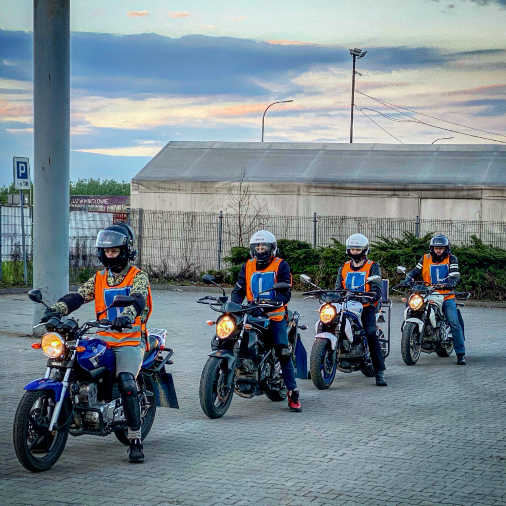 Czterech kursantów w kamizelkach z literą L siedzi na motocyklach Yamaha na placu manewrowym; w tle budynek magazynowy i słup z parkingiem dla TIR.
