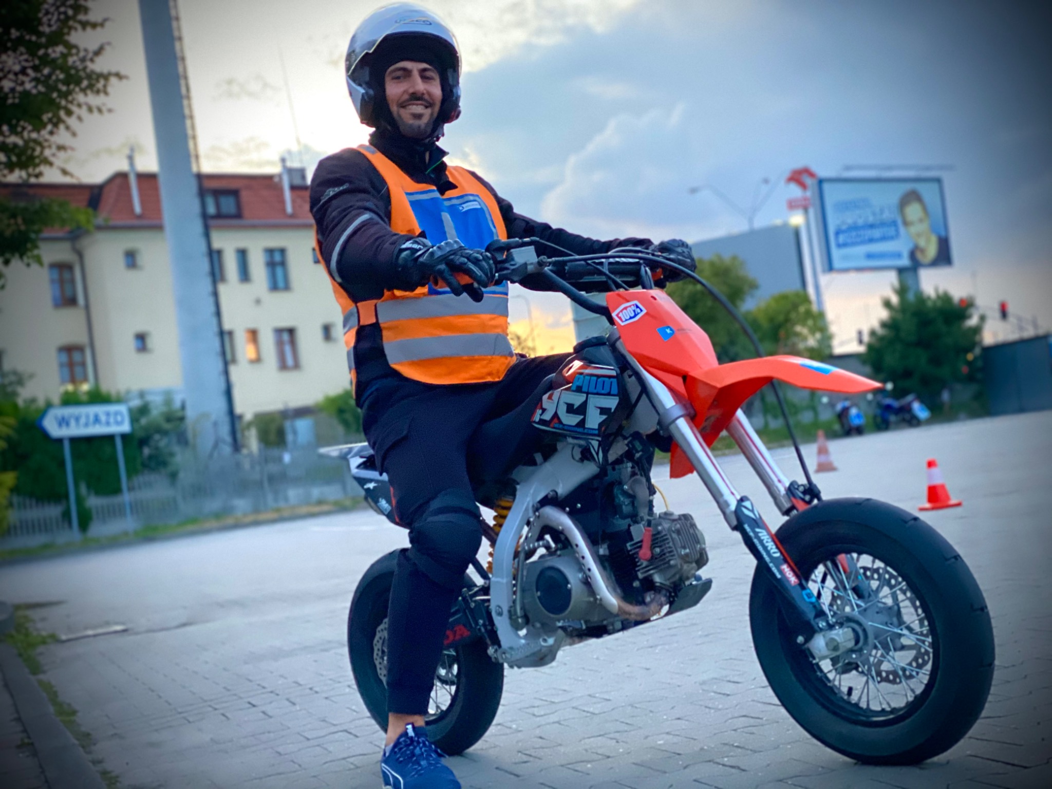 Uśmiechnięty instruktor w kasku i kamizelce odblaskowej siedzi na motocyklu typu supermoto z pomarańczowym błotnikiem na placu manewrowym, w tle budynek i billboard.