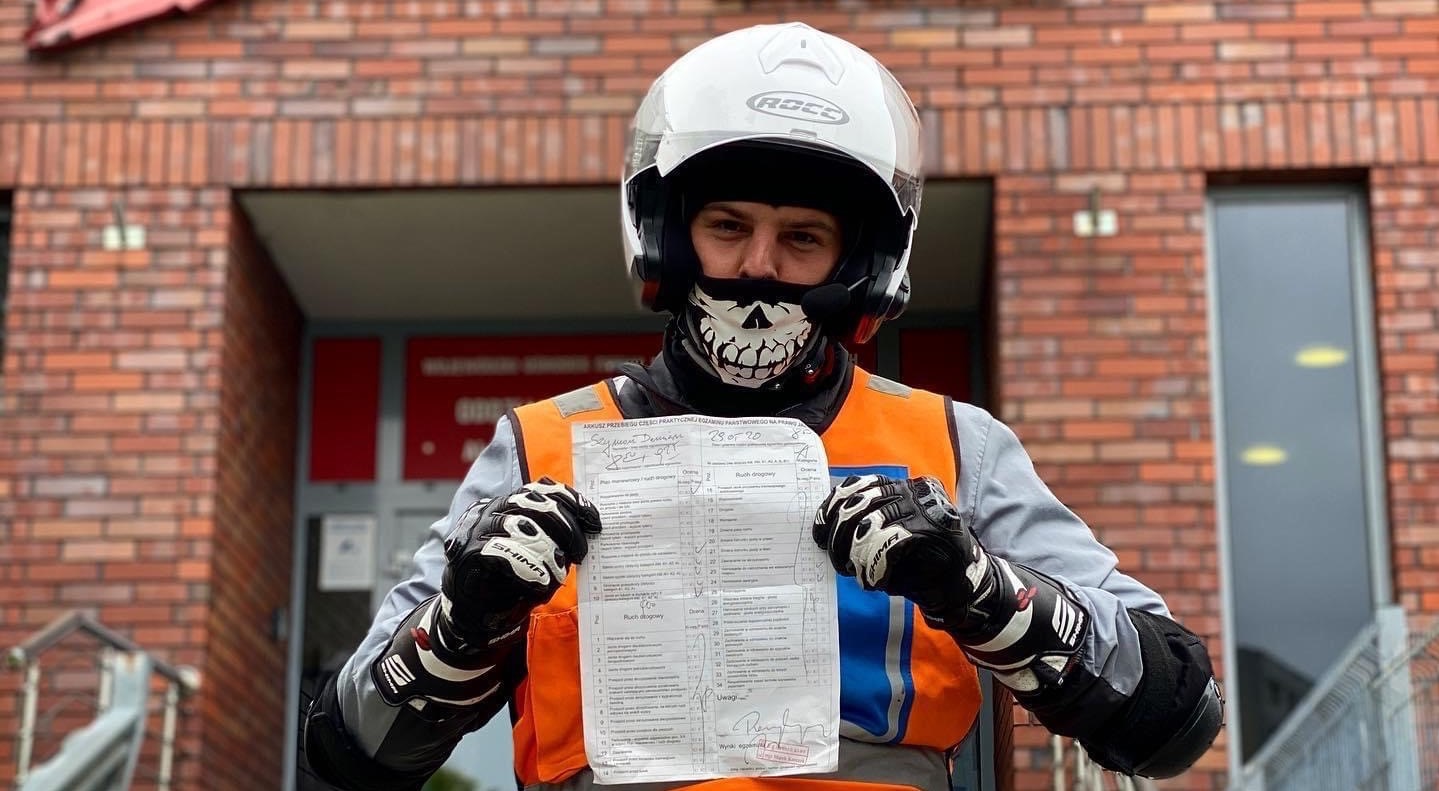 Egzaminator w kasku i masce z motywem czaszki, trzymający arkusz przebiegu egzaminu praktycznego na prawo jazdy, na tle budynku z cegły.