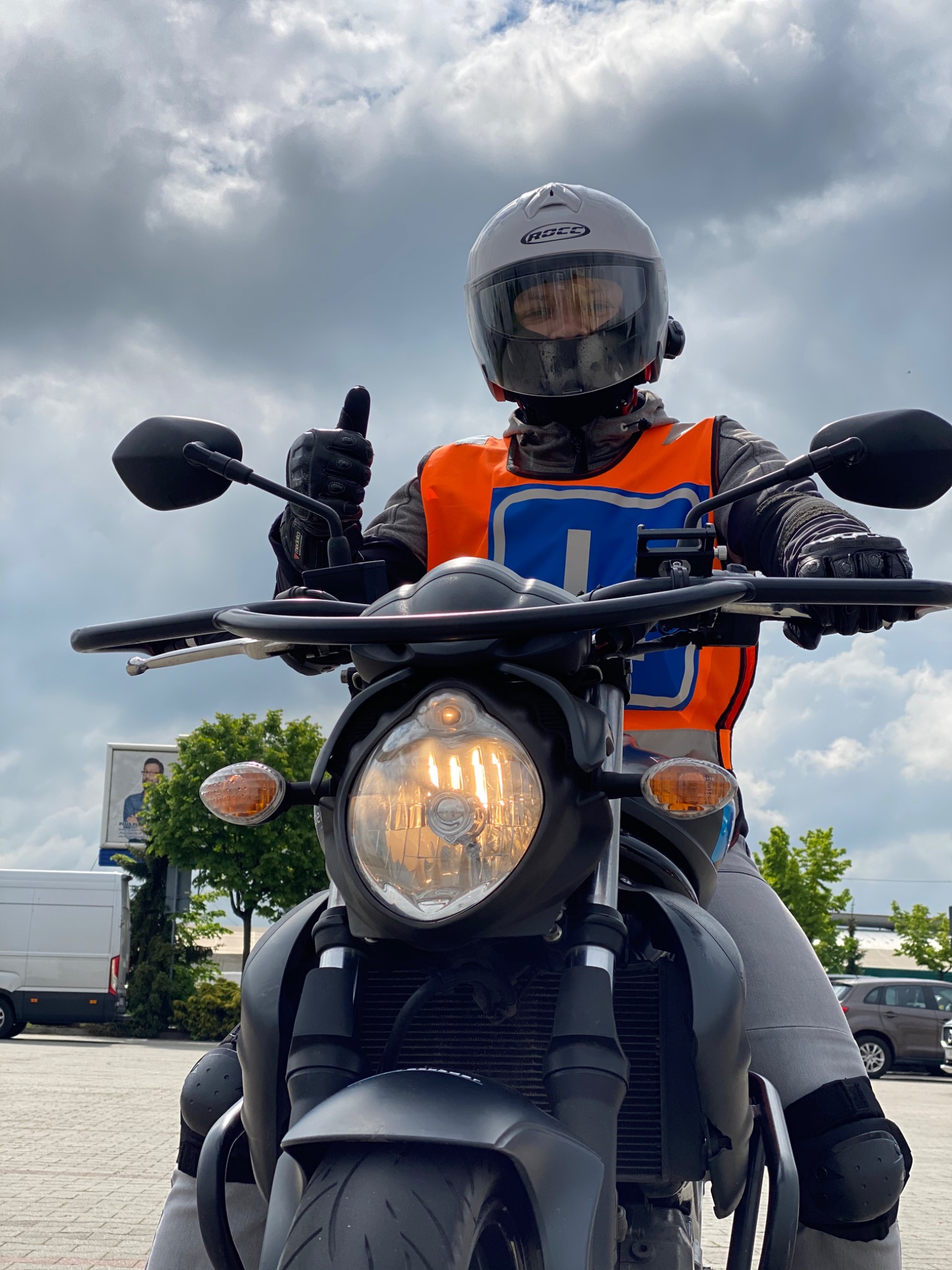 Instruktor szkoły jazdy motocyklem, w kasku i kamizelce odblaskowej, z podniesionym kciukiem na tle pochmurnego nieba.