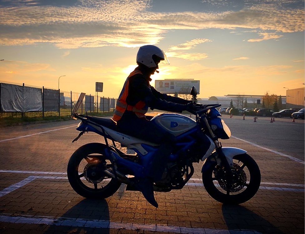 Sylwetka kursanta w kasku i kamizelce odblaskowej na motocyklu Suzuki SV650, na placu manewrowym o wschodzie słońca.