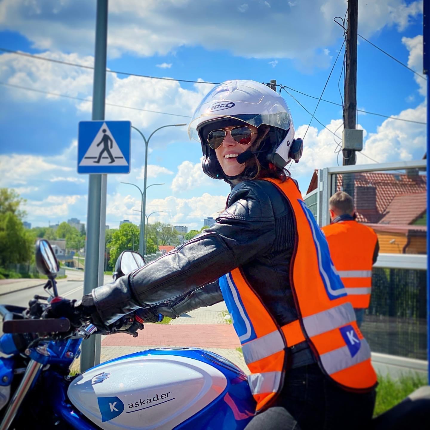 Uśmiechnięta kursantka w kasku i kamizelce odblaskowej na motocyklu z logo 'askader' na tle miejskiego krajobrazu i znaku przejścia dla pieszych.