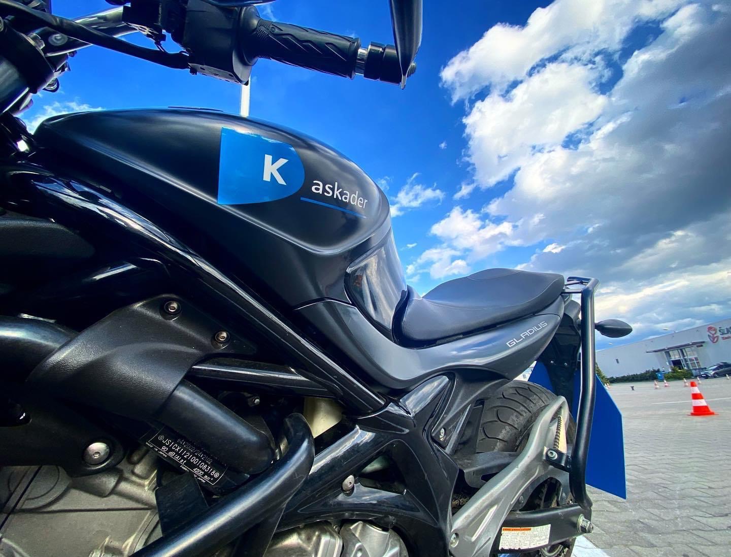 Czarny motocykl marki Gladius z niebieskim logo firmy 'Kaskader' na tle błękitnego nieba z chmurami i parkingu ze słupkami ostrzegawczymi.