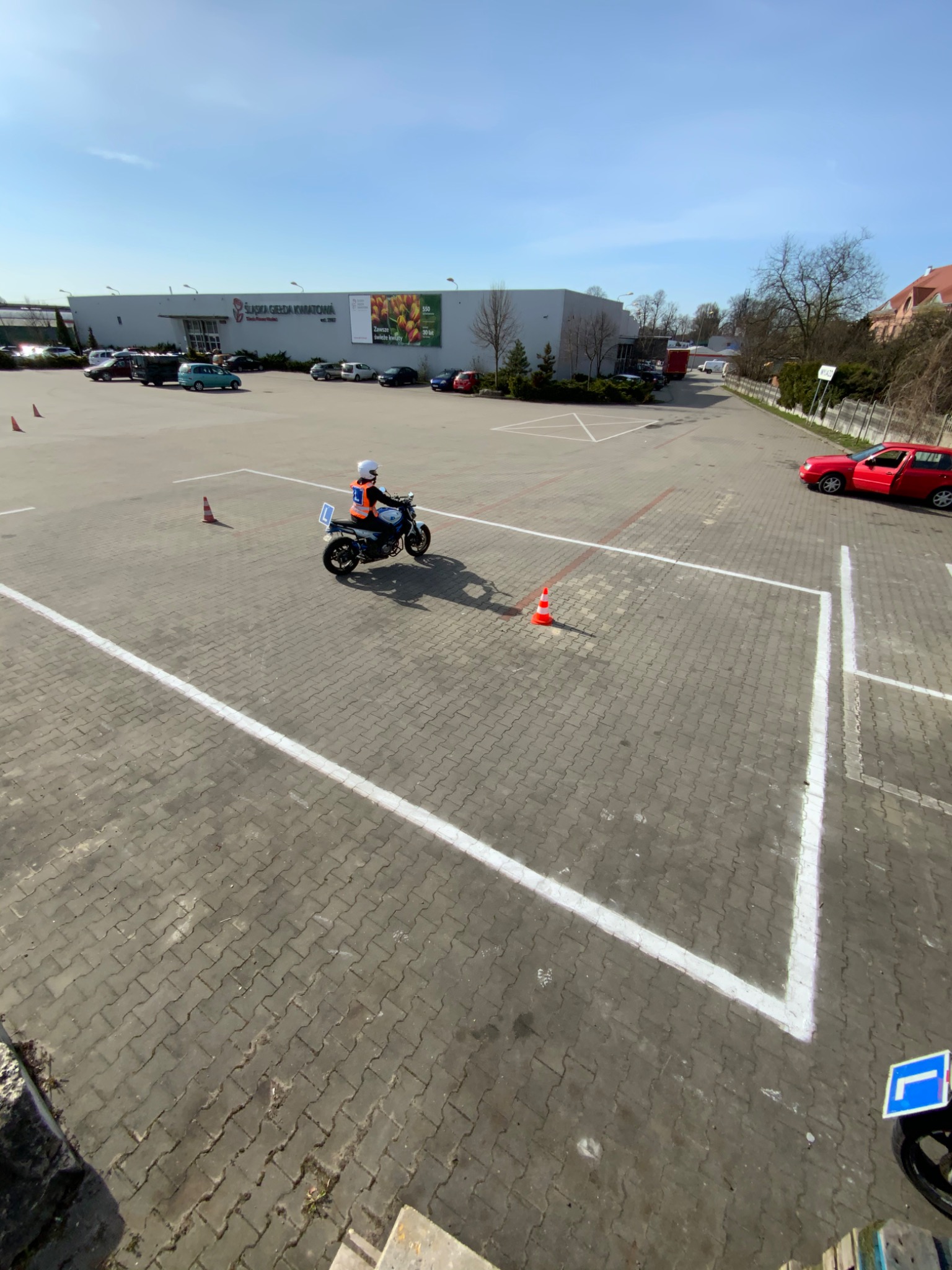 Osoba w kasku i kamizelce odblaskowej na motocyklu trójkołowym ćwiczy manewry na placu z wyznaczonymi liniami i pachołkami, w tle budynek handlowy i zaparkowane samochody.