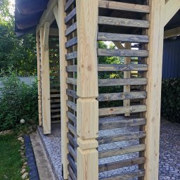 Marcin Skrętkowicz Usługi Ogólnobudowlane - Drewniana altana z jasnymi, surowymi słupami i poziomymi, ciemniejszymi listwami, otoczona zielenią ogrodu i kamienną nawierzchnią.