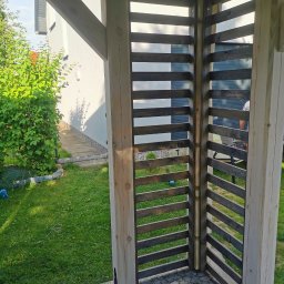 Marcin Skrętkowicz Usługi Ogólnobudowlane - Drewniana konstrukcja z poziomymi, ciemnymi listwami, tworząca ażurową ścianę, widoczna na tle zielonego trawnika i jasnego budynku.