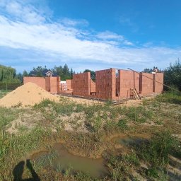 Marcin Skrętkowicz Usługi Ogólnobudowlane - Wznoszenie ścian parterowego domu z czerwonej cegły. Widoczny fragment konstrukcji, piasek i zarośnięty teren. Błękitne niebo z chmurami w tle.