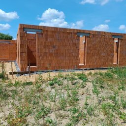 Marcin Skrętkowicz Usługi Ogólnobudowlane - Wznoszenie ścian parterowego domu z czerwonej cegły ceramicznej. Widoczne otwory okienne i drzwiowe. Prace murarskie w toku, teren budowy z roślinnością.