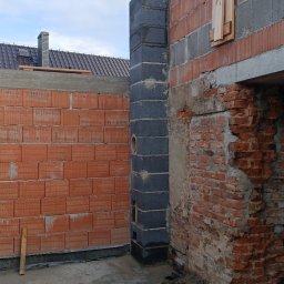 Marcin Skrętkowicz Usługi Ogólnobudowlane - Murowanie komina z bloczków w Nysie. Widoczne cegły, zaprawa w wiadrze i fragment dachu. Prace murarskie w toku, zarys budynku w stanie surowym.