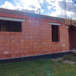 Marcin Skrętkowicz Usługi Ogólnobudowlane - Budowa domu jednorodzinnego w trakcie wznoszenia ścian z czerwonej cegły. Widoczne otwory okienne i drzwiowe, szalunki, zbrojenia oraz rusztowania.