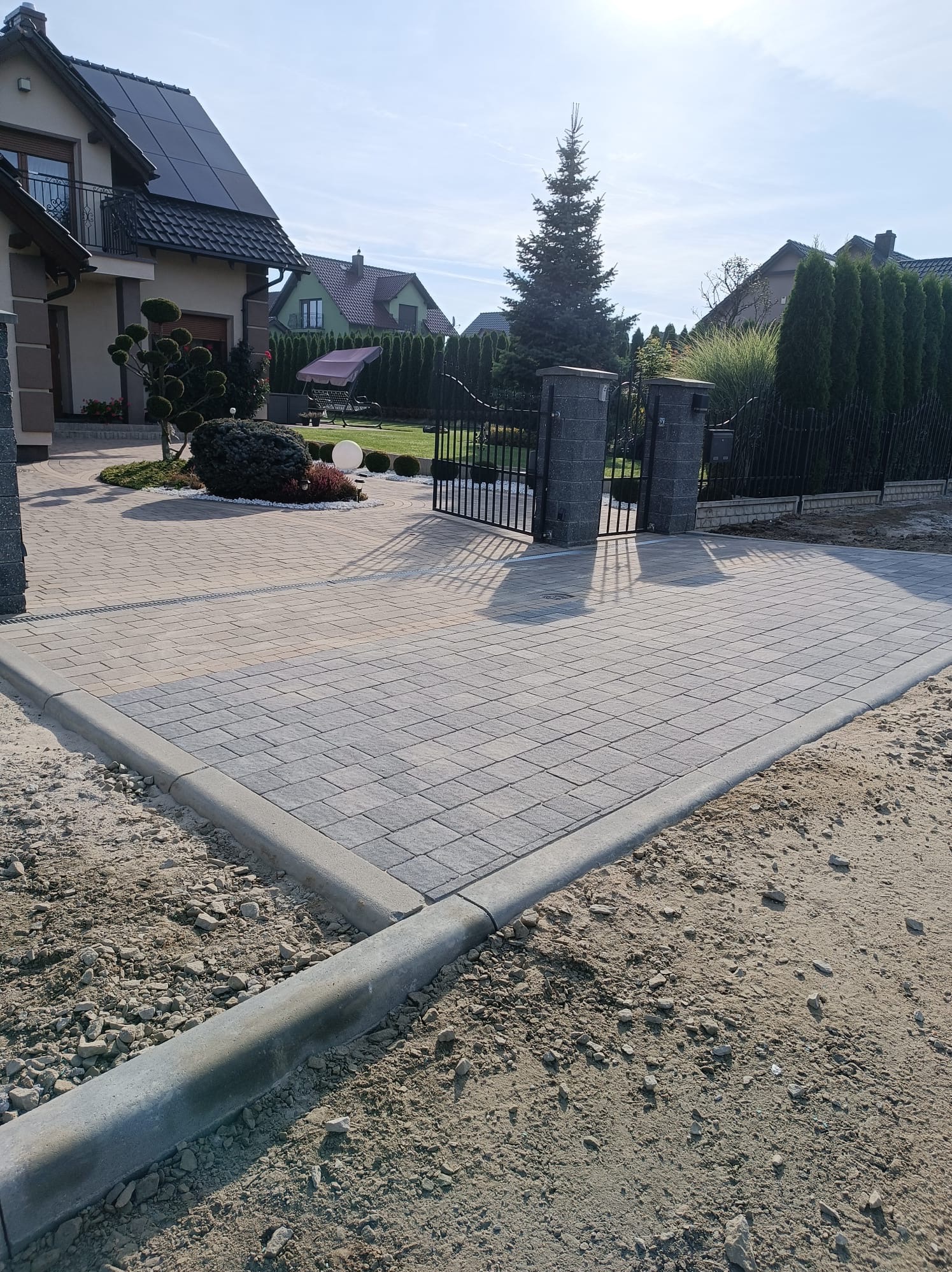 Nowa, szara kostka brukowa ułożona na podjeździe z widoczną bramą wjazdową z metalowymi prętami, otoczona zadbanym ogrodem z tujami i kulistymi krzewami.