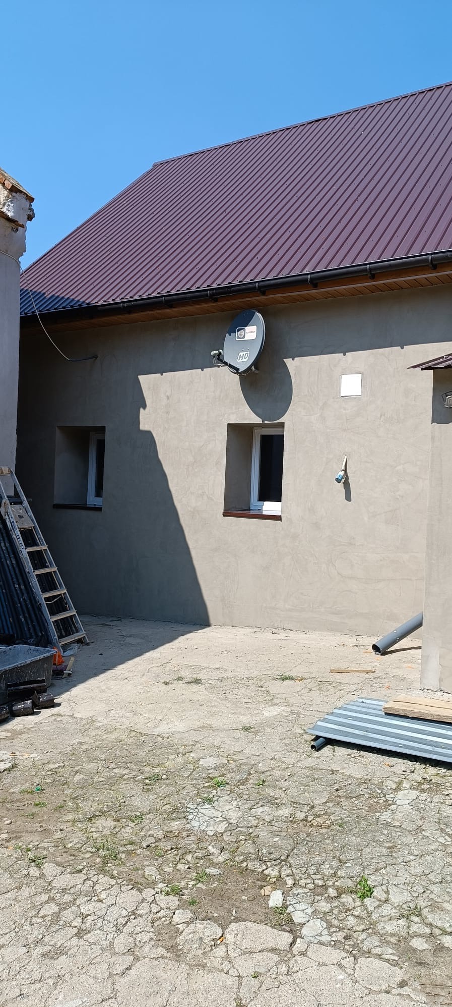 Świeżo otynkowana ściana budynku z zamontowaną anteną satelitarną, widoczne okna z brązowymi ramami i fragment dachu z metalowej blachy w kolorze bordowym, drabina oparta o ścianę.