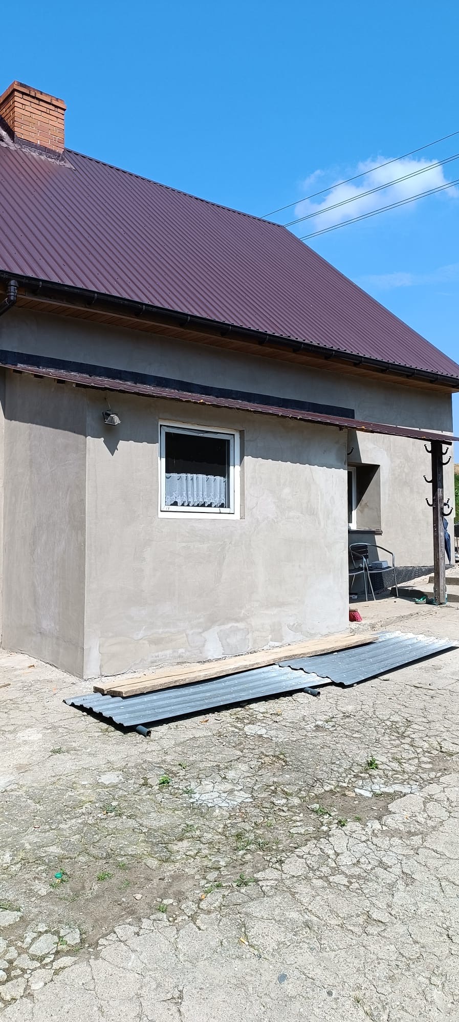 Dom z szarą elewacją i brązowym dachem z blachy falistej, przygotowany do prac remontowych, z widocznymi deskami i arkuszami blachy na ziemi.