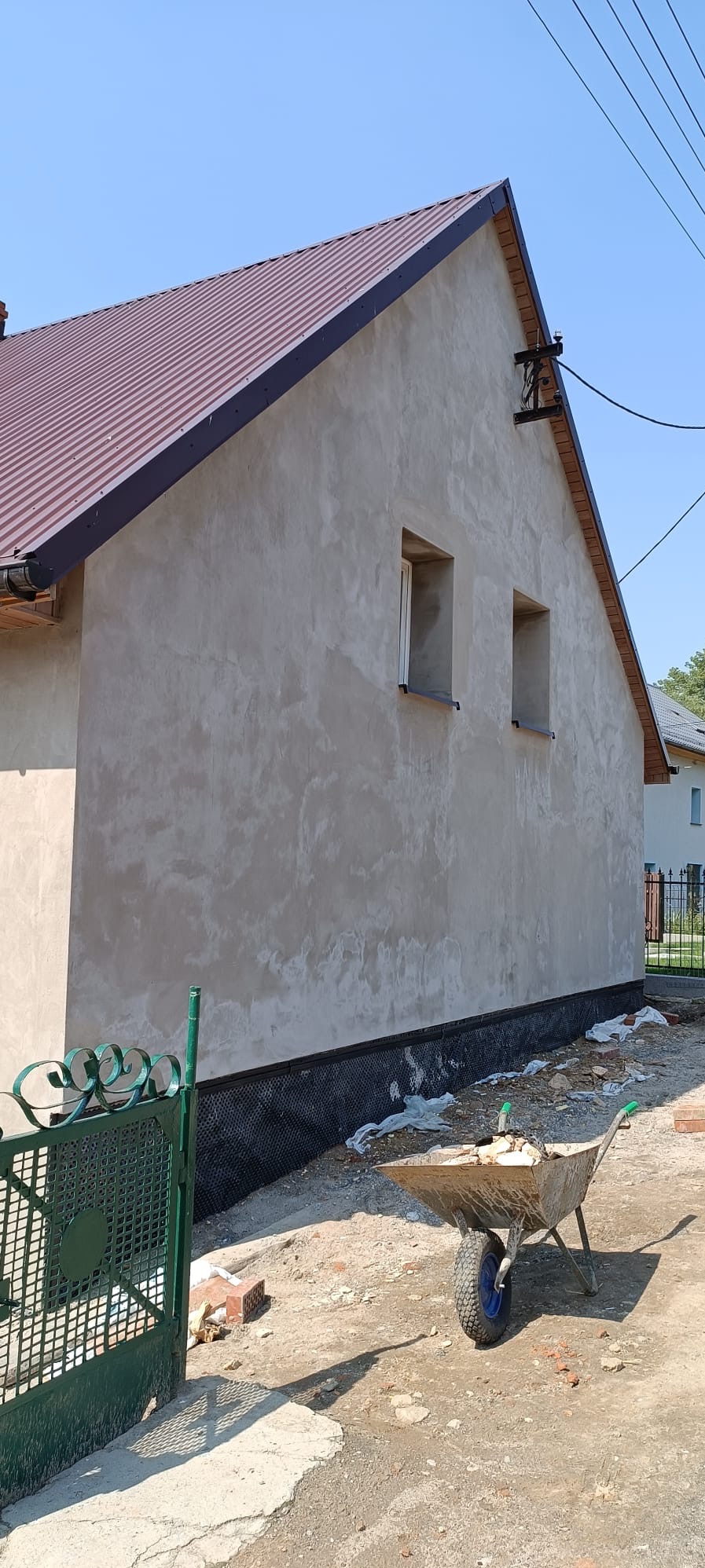 Ściana domu w trakcie renowacji elewacji, z widoczną surową zaprawą, dwoma małymi oknami i fragmentem dachu z blachy. W tle widoczny fragment ogrodzenia i sąsiedni budynek, a na pierwszym planie...