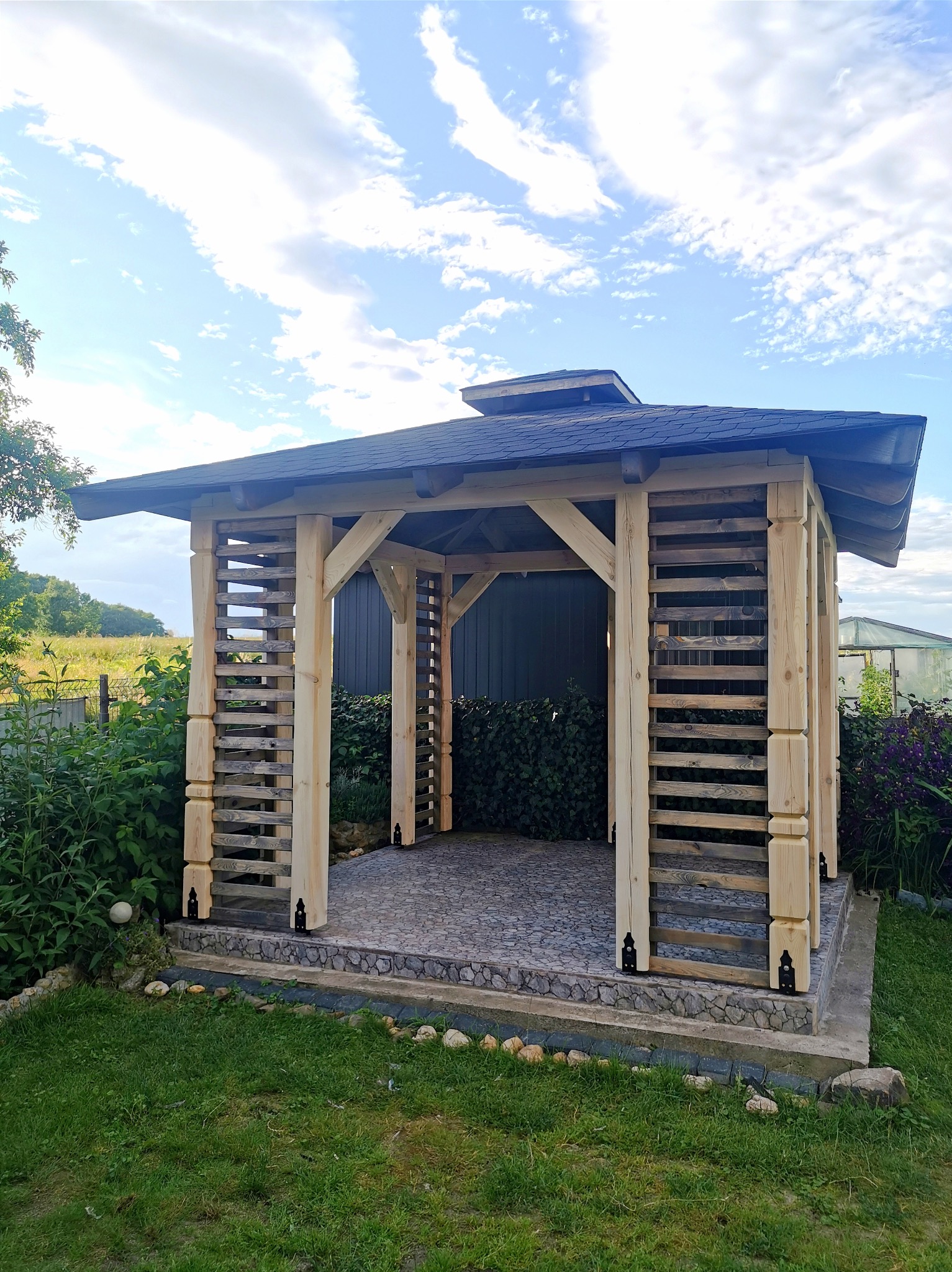 Drewniana altana ogrodowa z ażurowymi ścianami w Nysie, wykończona ciemną dachówką, otoczona zielenią i kamieniami.