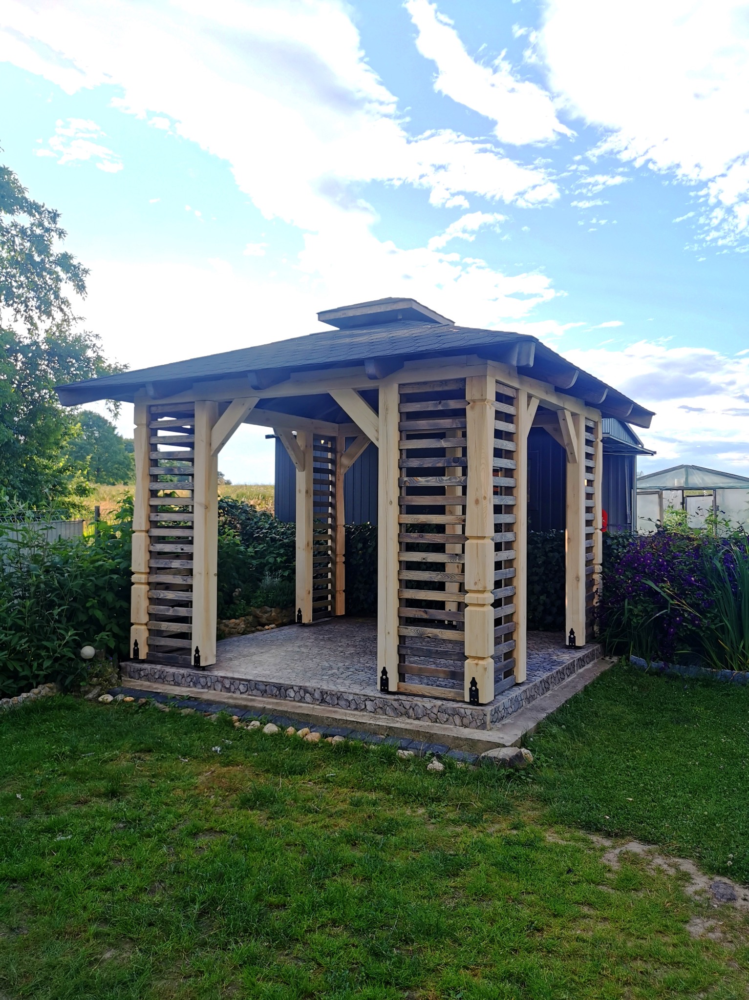 Drewniana altana ogrodowa z ażurowymi ścianami, pokryta ciemną dachówką, posadowiona na kamiennej podłodze, otoczona zielenią i kwiatami w słoneczny dzień.