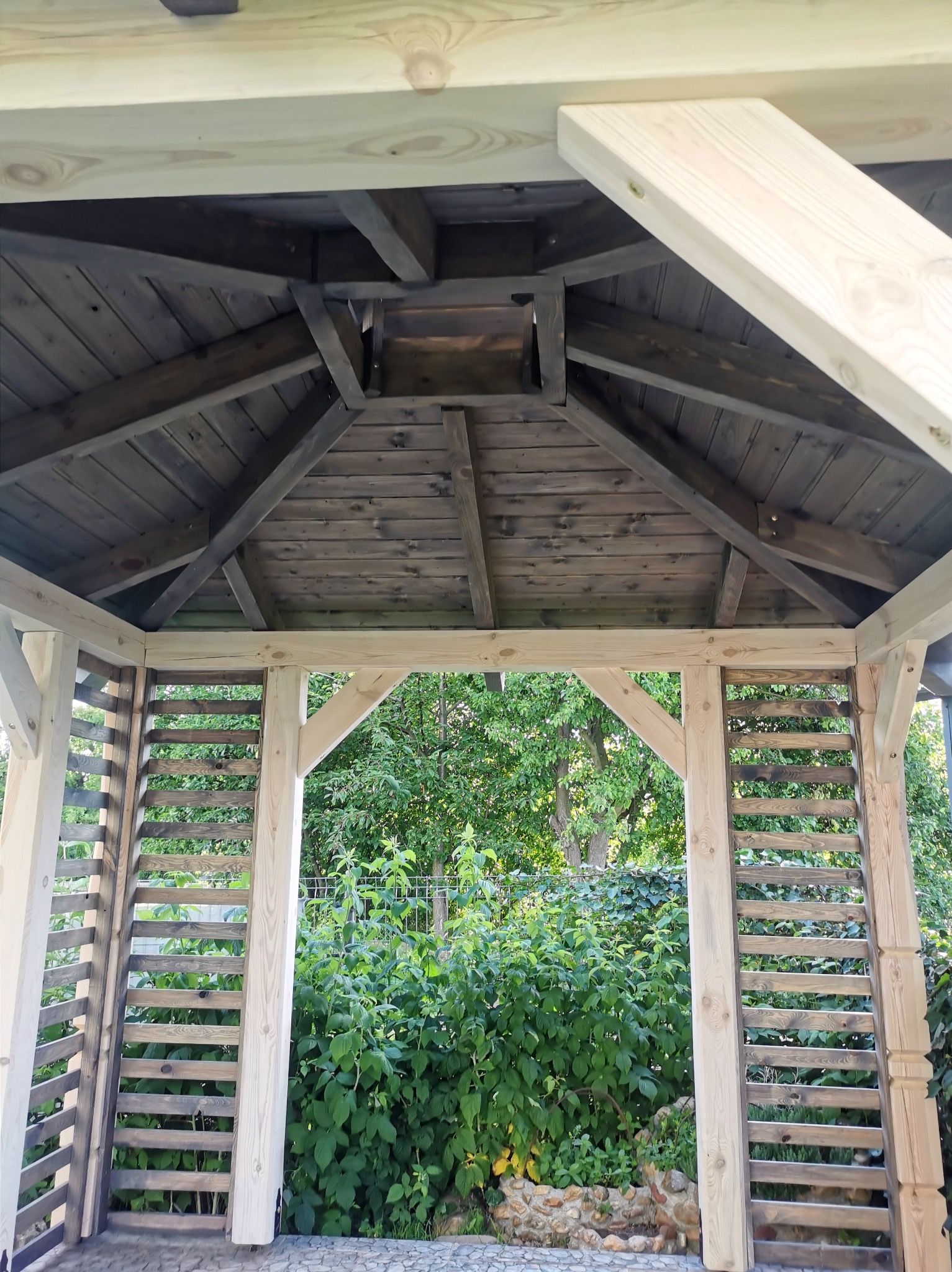 Wnętrze drewnianej altany z widokiem na zieleń ogrodu, konstrukcja dachu z belek w odcieniach brązu i szarości, jasne drewniane słupy i ażurowe panele boczne.