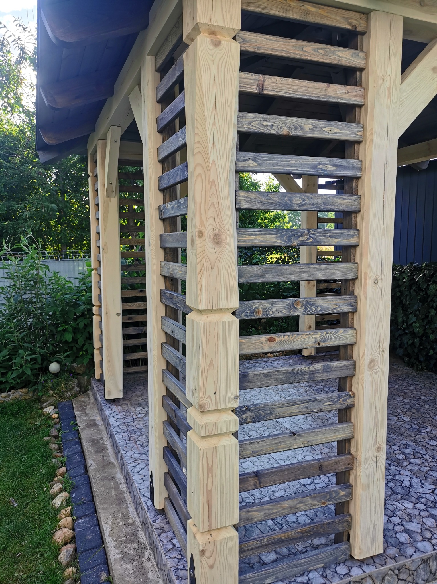 Drewniana altana z jasnymi, surowymi słupami i poziomymi, ciemniejszymi listwami, otoczona zielenią ogrodu i kamienną nawierzchnią.