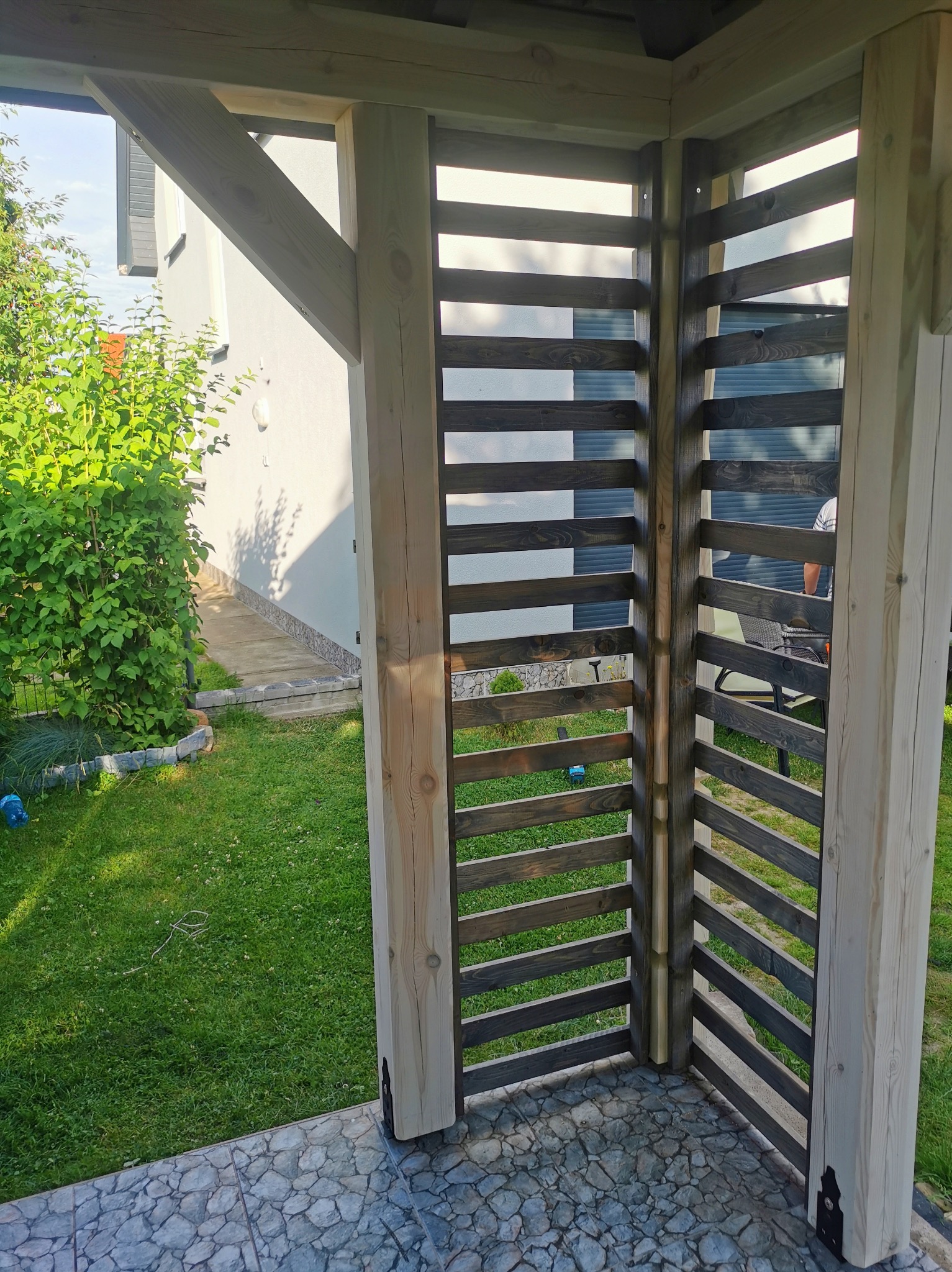 Drewniana konstrukcja z poziomymi, ciemnymi listwami, tworząca ażurową ścianę, widoczna na tle zielonego trawnika i jasnego budynku.