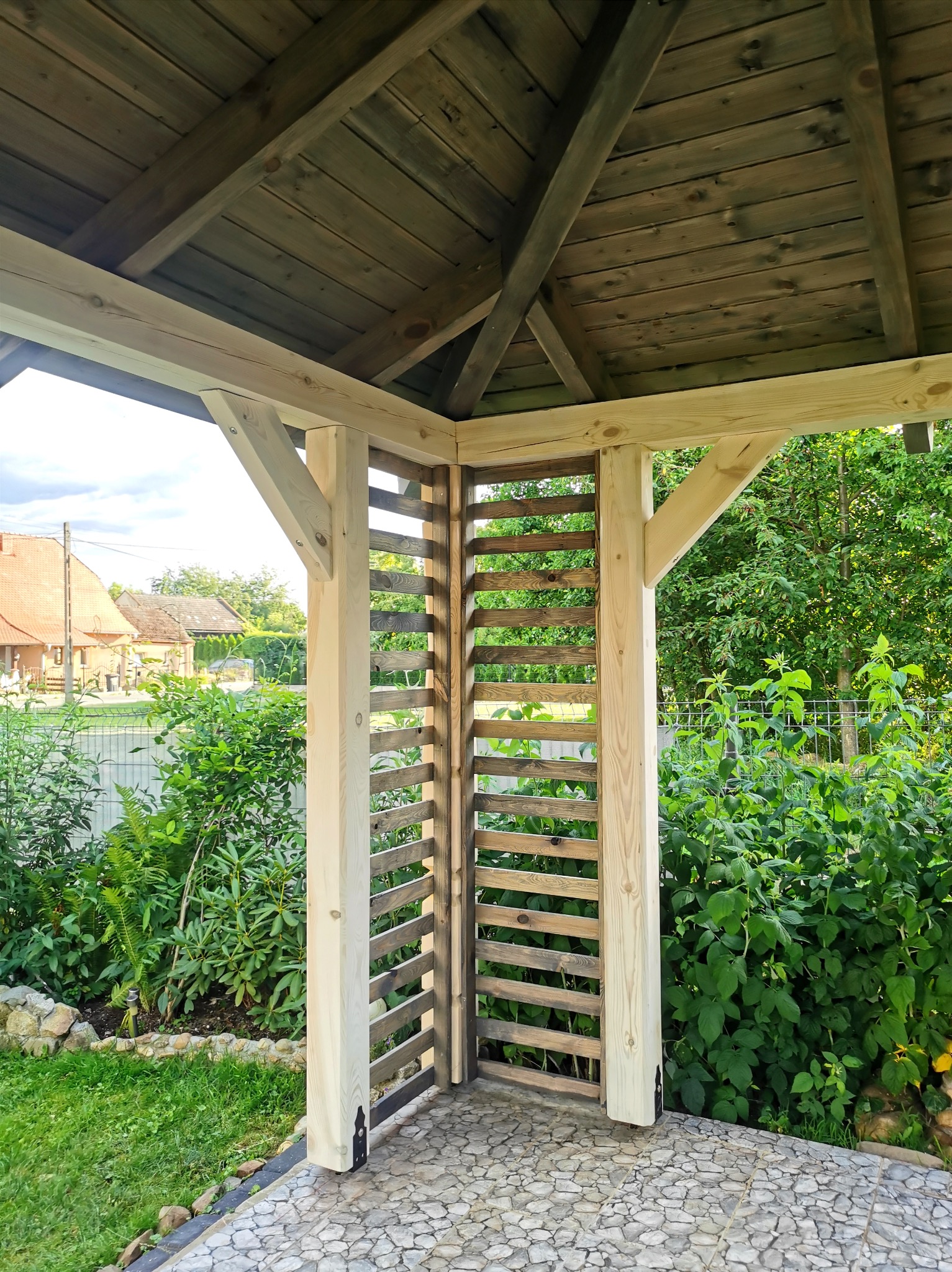 Drewniana konstrukcja altany z ażurowym panelem bocznym, widok na ogród i zabudowania w tle.
