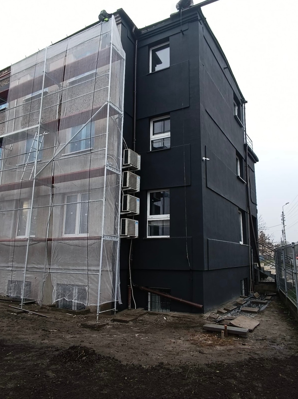 Budynek wielorodzinny w trakcie prac elewacyjnych: jedna ściana w rusztowaniach osłoniętych folią, druga pomalowana na czarno, widoczne klimatyzatory.