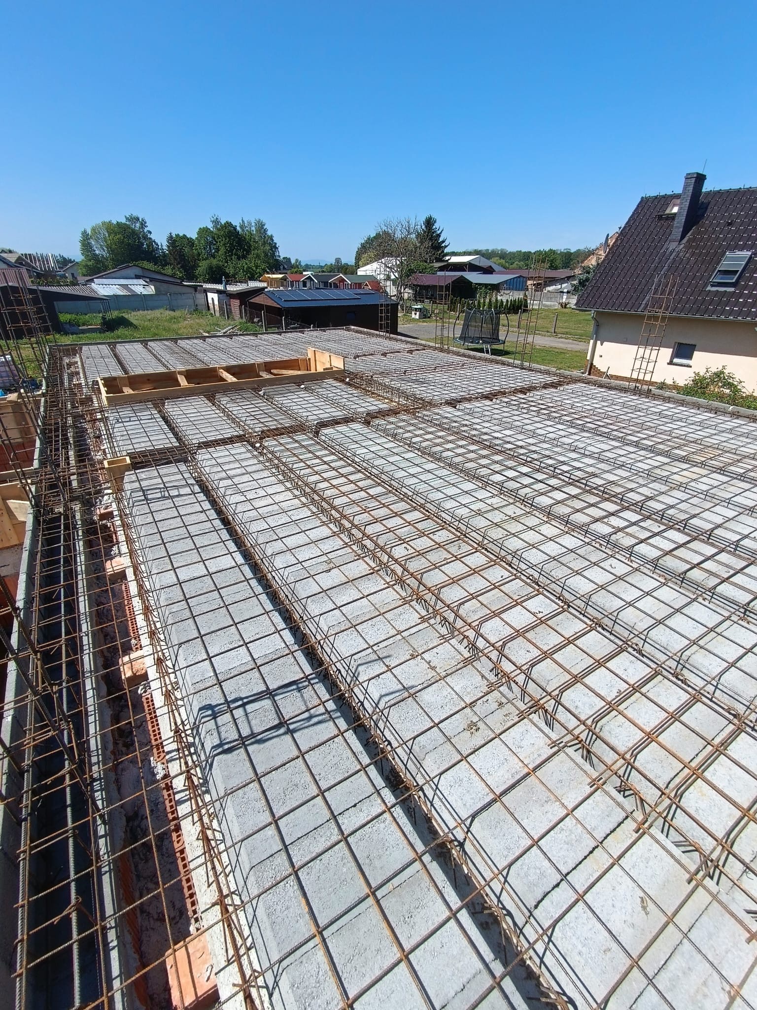 Zbrojenie stropu budynku jednorodzinnego z betonu komórkowego, widok z góry na siatkę z prętów stalowych i pustaki szalunkowe. W tle zabudowa jednorodzinna i błękitne niebo.