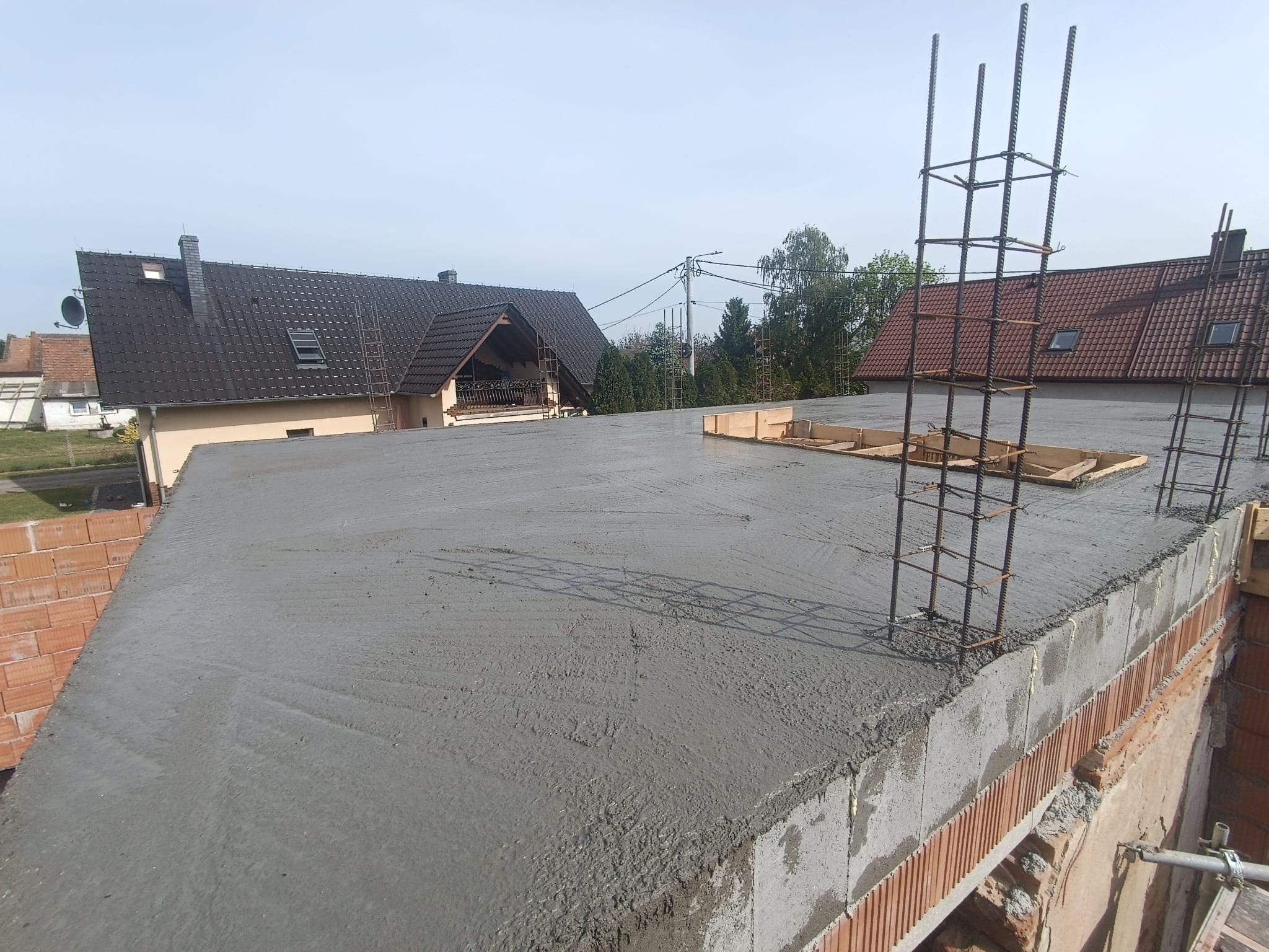 Świeżo wylany beton na stropie budynku z widocznym zbrojeniem i szalunkiem, w tle zabudowa jednorodzinna w słoneczny dzień. Prace budowlane w toku.
