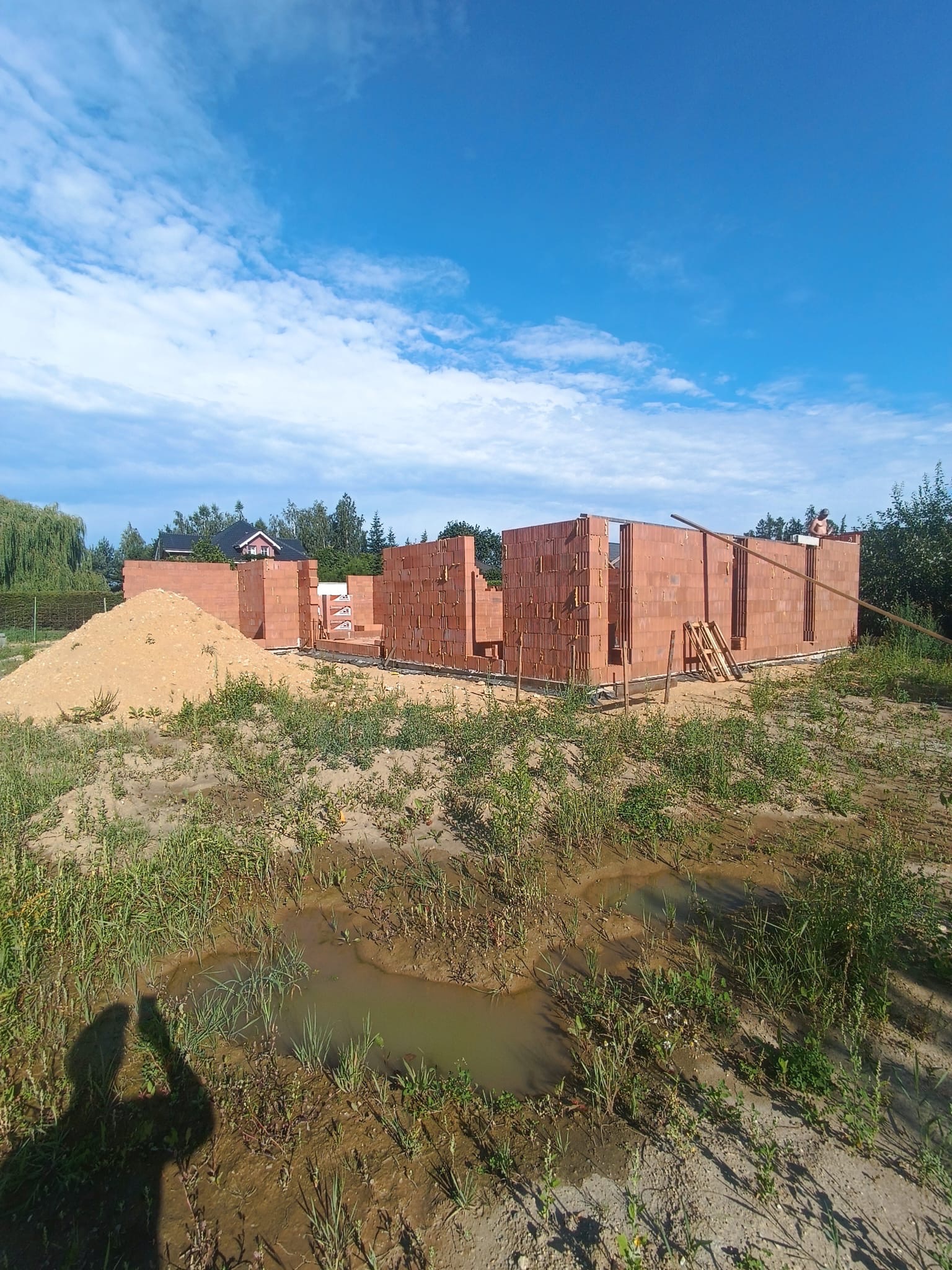 Wznoszenie ścian parterowego domu z czerwonej cegły. Widoczny fragment konstrukcji, piasek i zarośnięty teren. Błękitne niebo z chmurami w tle.