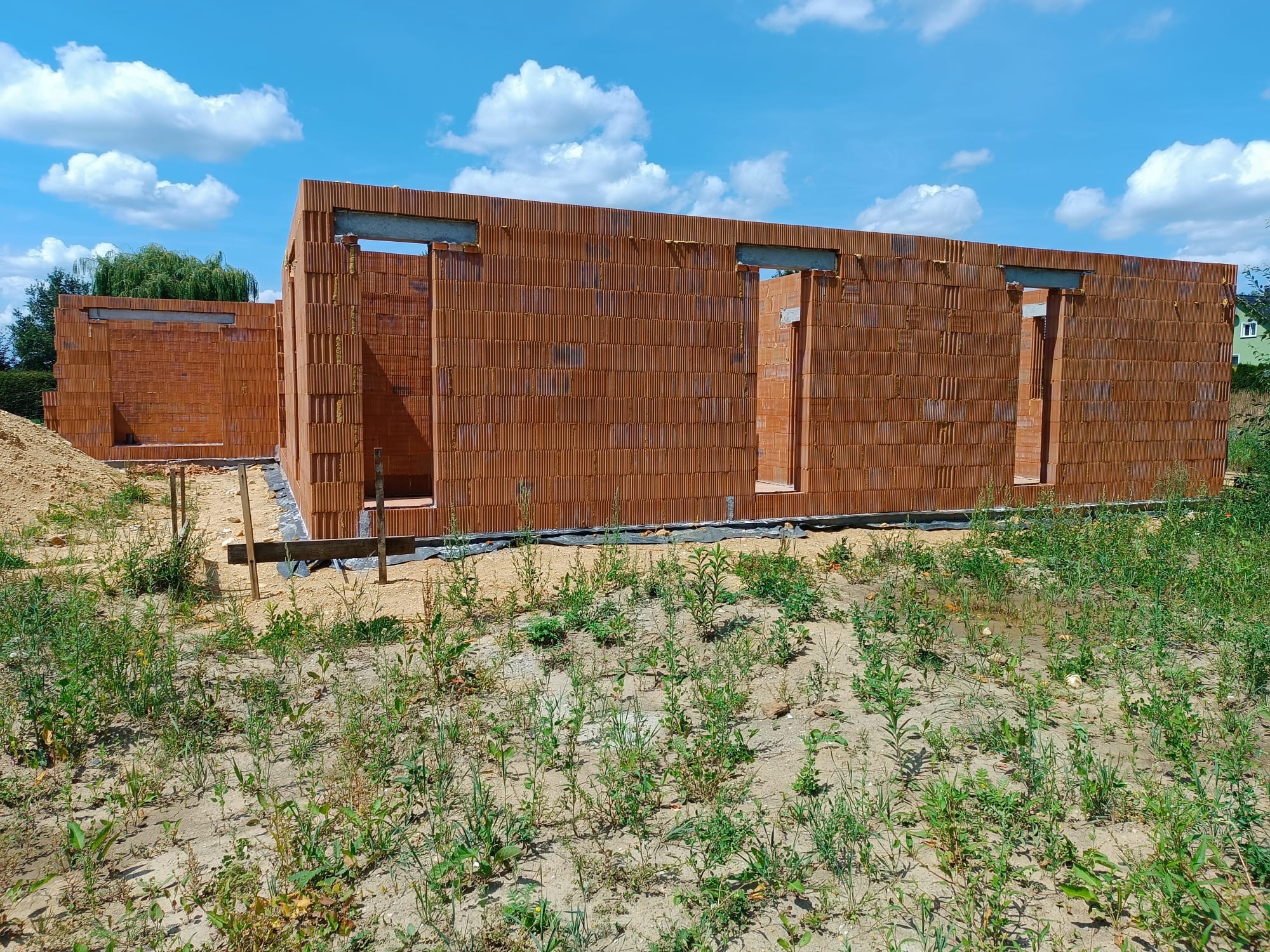 Wznoszenie ścian parterowego domu z czerwonej cegły ceramicznej. Widoczne otwory okienne i drzwiowe. Prace murarskie w toku, teren budowy z roślinnością.