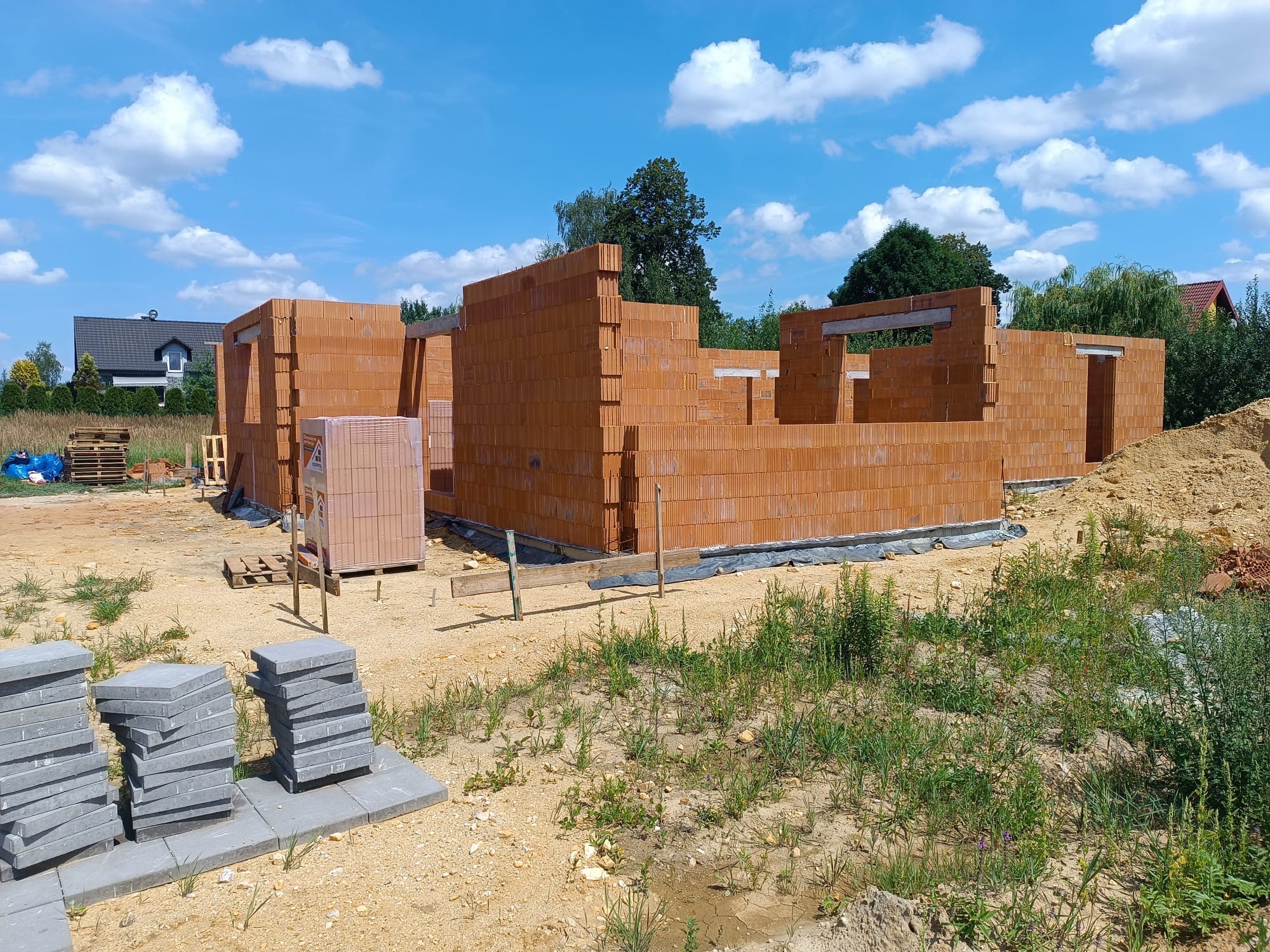 Budowa domu jednorodzinnego w trakcie wznoszenia ścian z czerwonej cegły. Widoczne palety z cegłami i ułożone płyty chodnikowe. Błękitne niebo z białymi chmurami w tle.