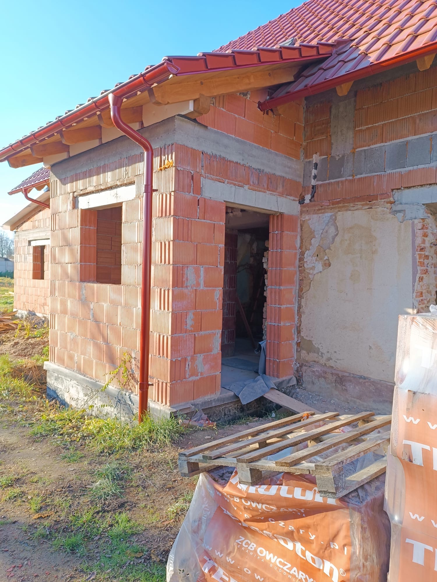 Budowa domu w trakcie realizacji: widoczne ściany z czerwonej cegły, okna bez szyb, czerwone rynny i paleta z materiałami budowlanymi na zewnątrz.