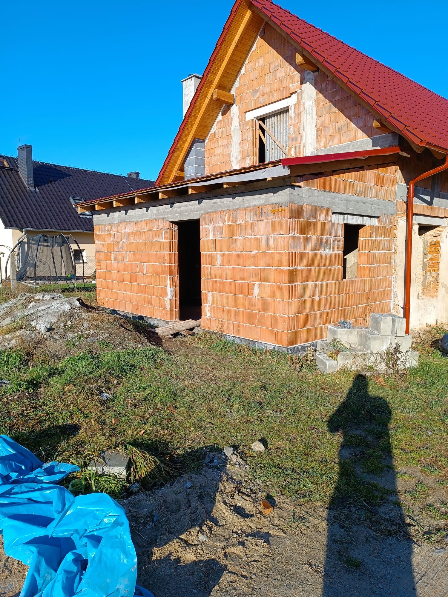 Budowa domu w stanie surowym otwartym: ściany z cegły ceramicznej, czerwony dach, widoczne elementy konstrukcyjne. Na pierwszym planie cień osoby.