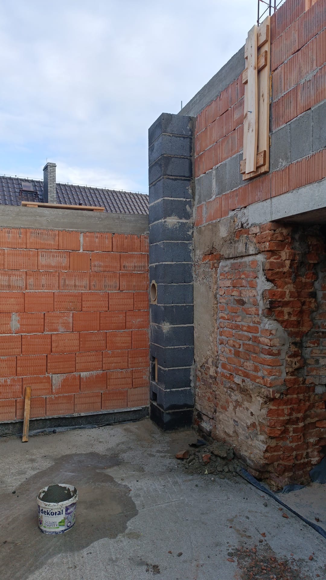 Murowanie komina z bloczków w Nysie. Widoczne cegły, zaprawa w wiadrze i fragment dachu. Prace murarskie w toku, zarys budynku w stanie surowym.