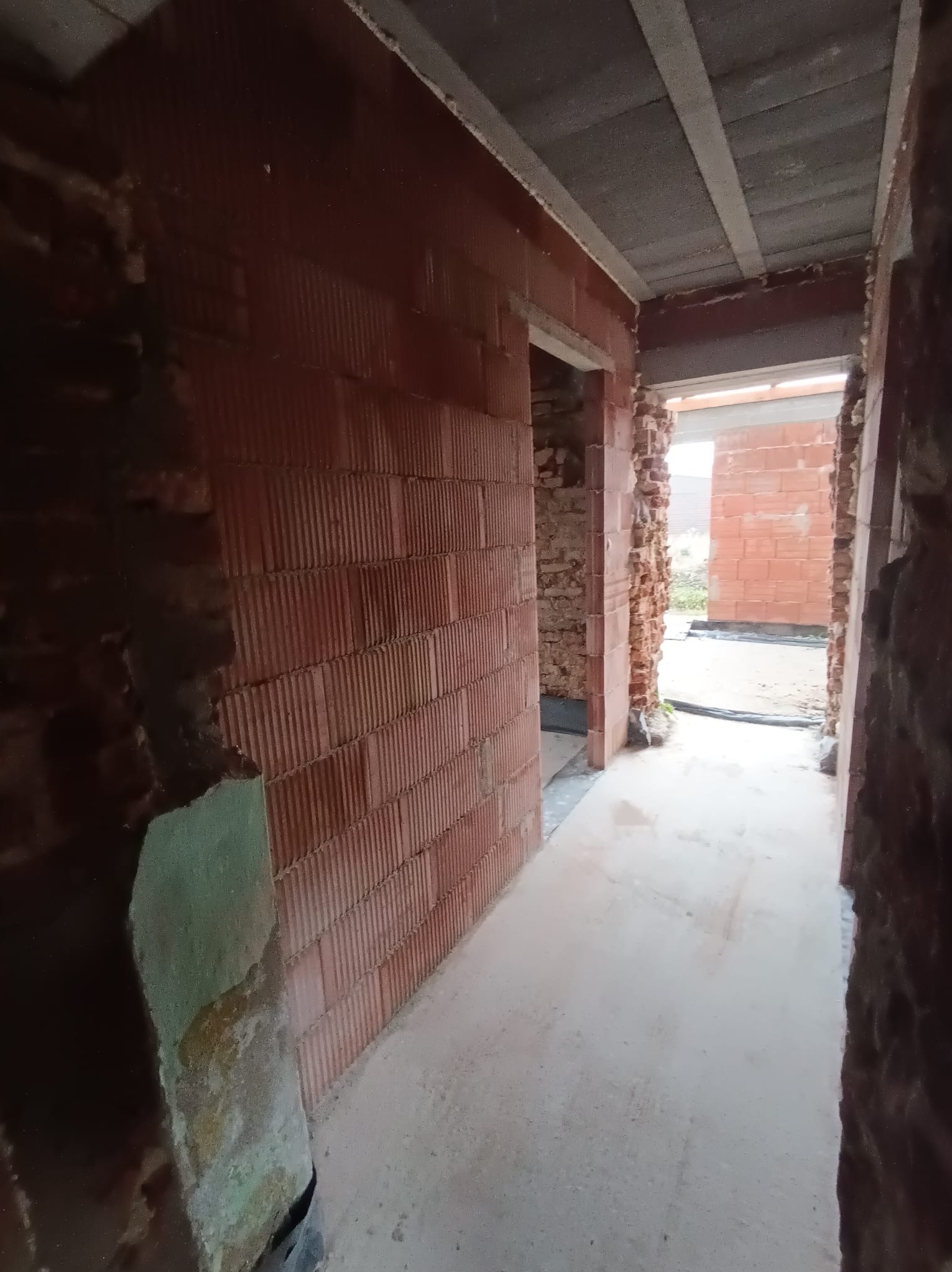 Wnętrze w trakcie budowy: ściany z czerwonej cegły dziurawki, surowy betonowy sufit, widok na zewnątrz przez otwór drzwiowy. Surowy stan deweloperski.