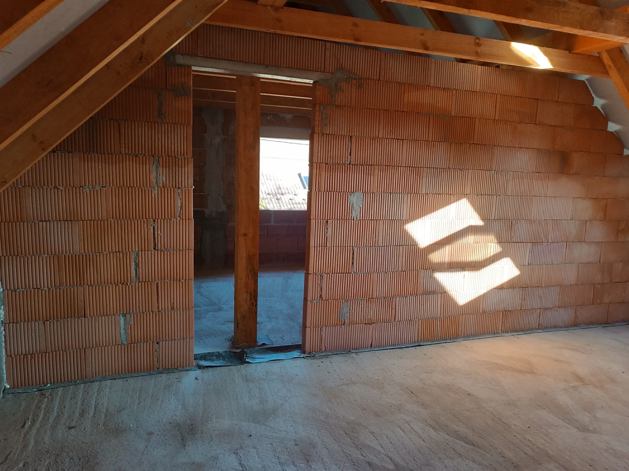 Wnętrze poddasza w trakcie budowy: ściana z czerwonej cegły dziurawki, drewniana konstrukcja dachu, widoczne fragmenty surowego betonu na podłodze i w tle.