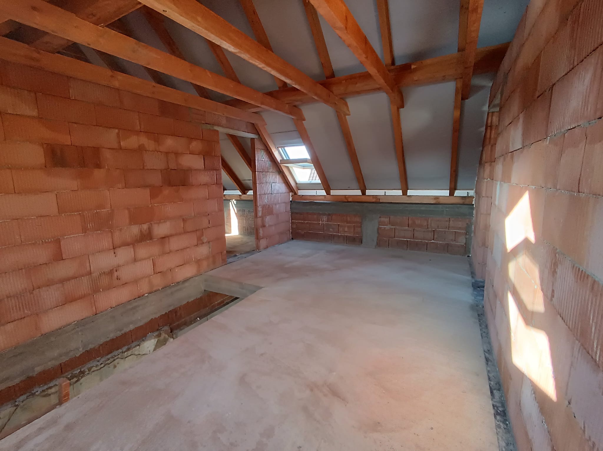 Surowy stan poddasza z oknem dachowym, ściany z czerwonej cegły, drewniana konstrukcja dachu, wylewka podłogowa. Widok na sąsiednie pomieszczenie.