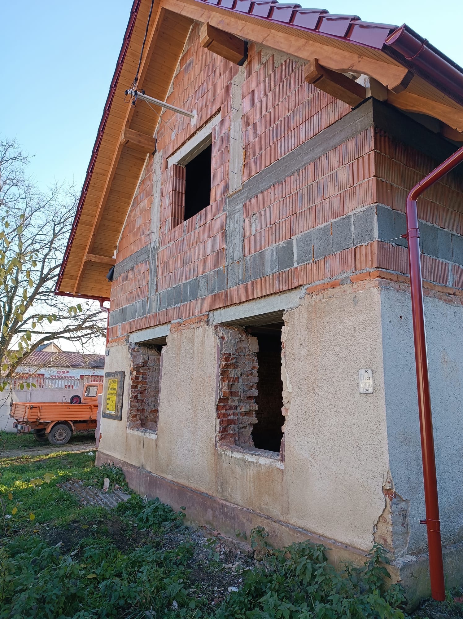 Budowa domu z czerwonej cegły, widoczne otwory okienne i fragment dachu z czerwoną dachówką. Na drugim planie drzewo i pomarańczowy samochód dostawczy.