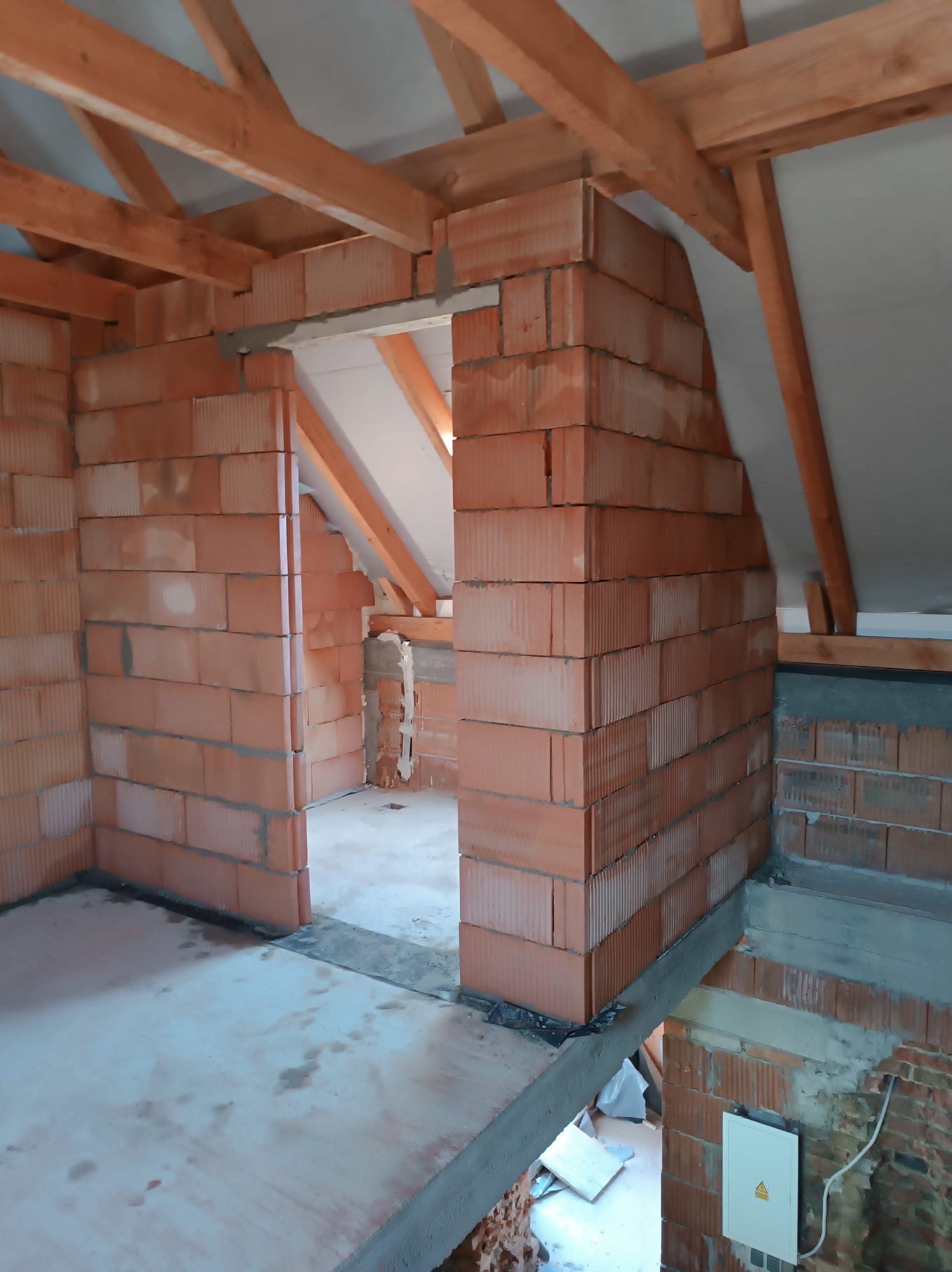 Wnętrze poddasza w trakcie budowy: ściany z czerwonej cegły, drewniana konstrukcja dachu, widoczny strop. Etap murowania ścian działowych.