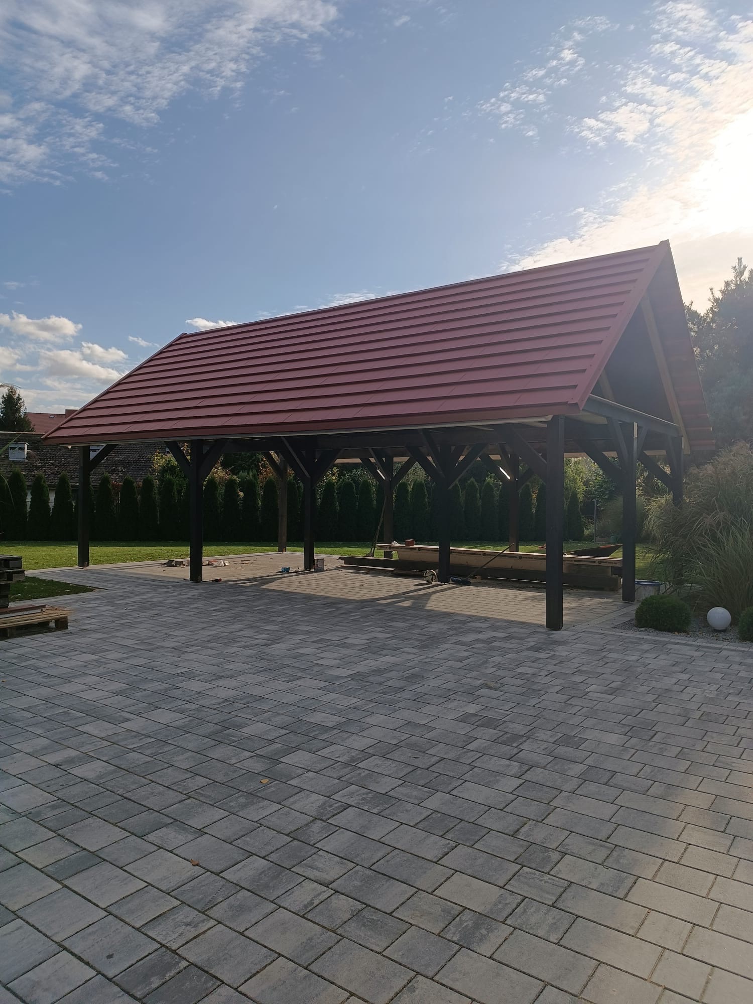 Zadaszenie tarasowe z ciemnego drewna i czerwoną dachówką, widok z zewnątrz na tle nieba i zieleni. Kostka brukowa na pierwszym planie.
