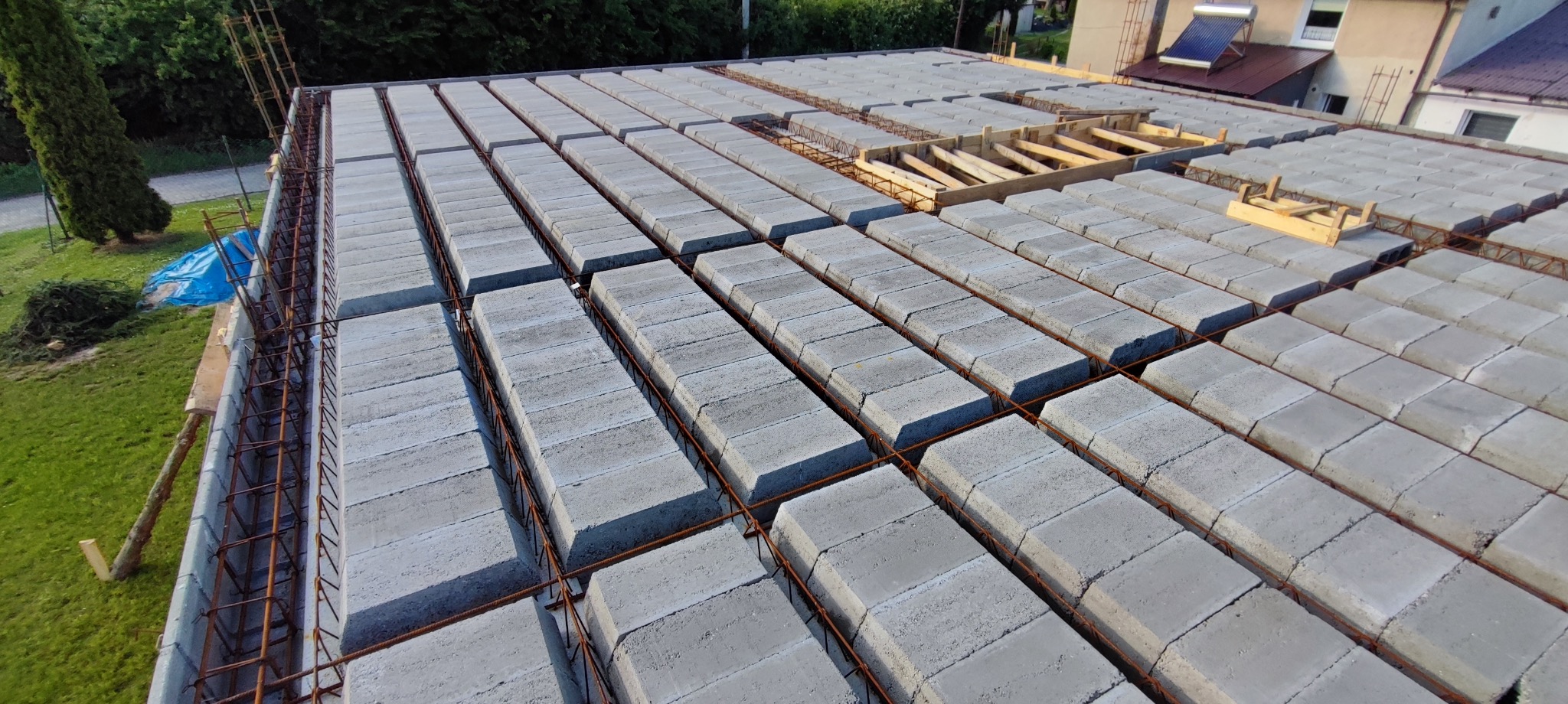 Zalany strop Teriva z szalunkiem obwodowym i zbrojeniem, widok z góry. Elementy drewniane w centralnej części. W tle budynek mieszkalny i zieleń.