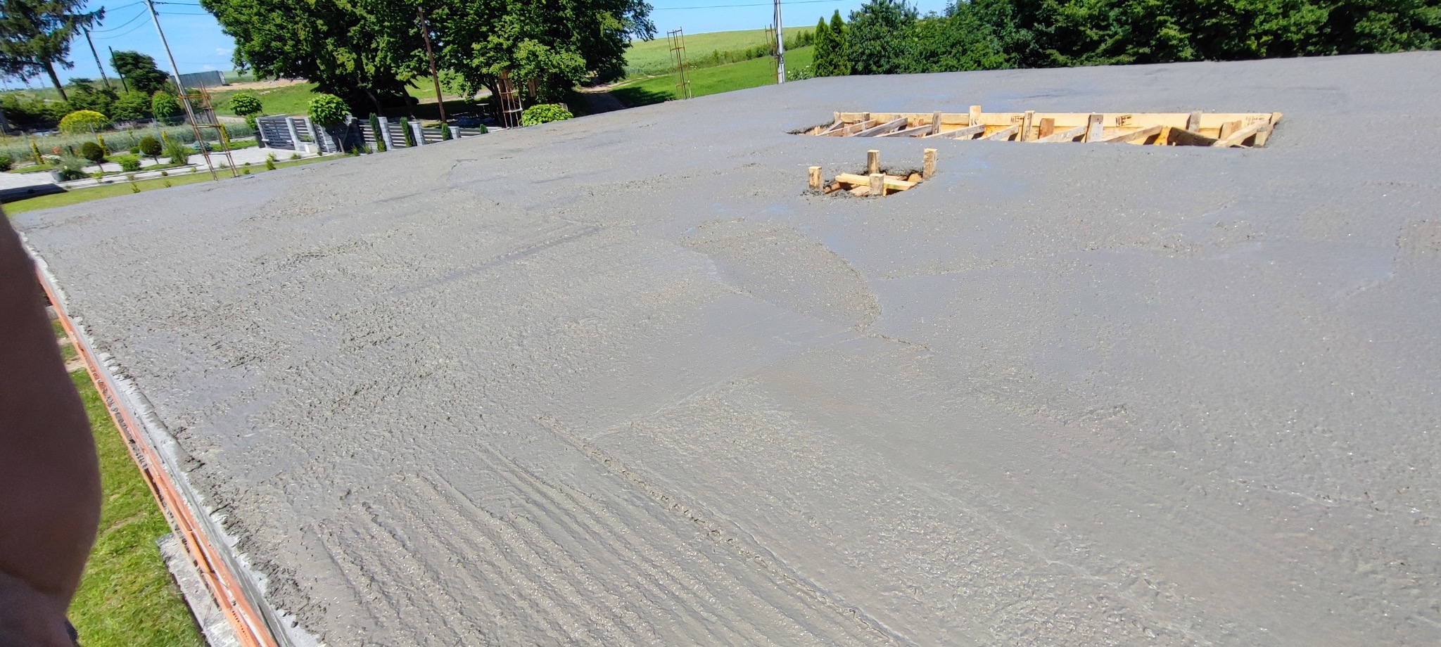 Świeżo wylany, gładki beton na dużej powierzchni z drewnianymi szalunkami w kilku miejscach. Widoczne ślady po rozprowadzaniu betonu, zielone otoczenie w tle.