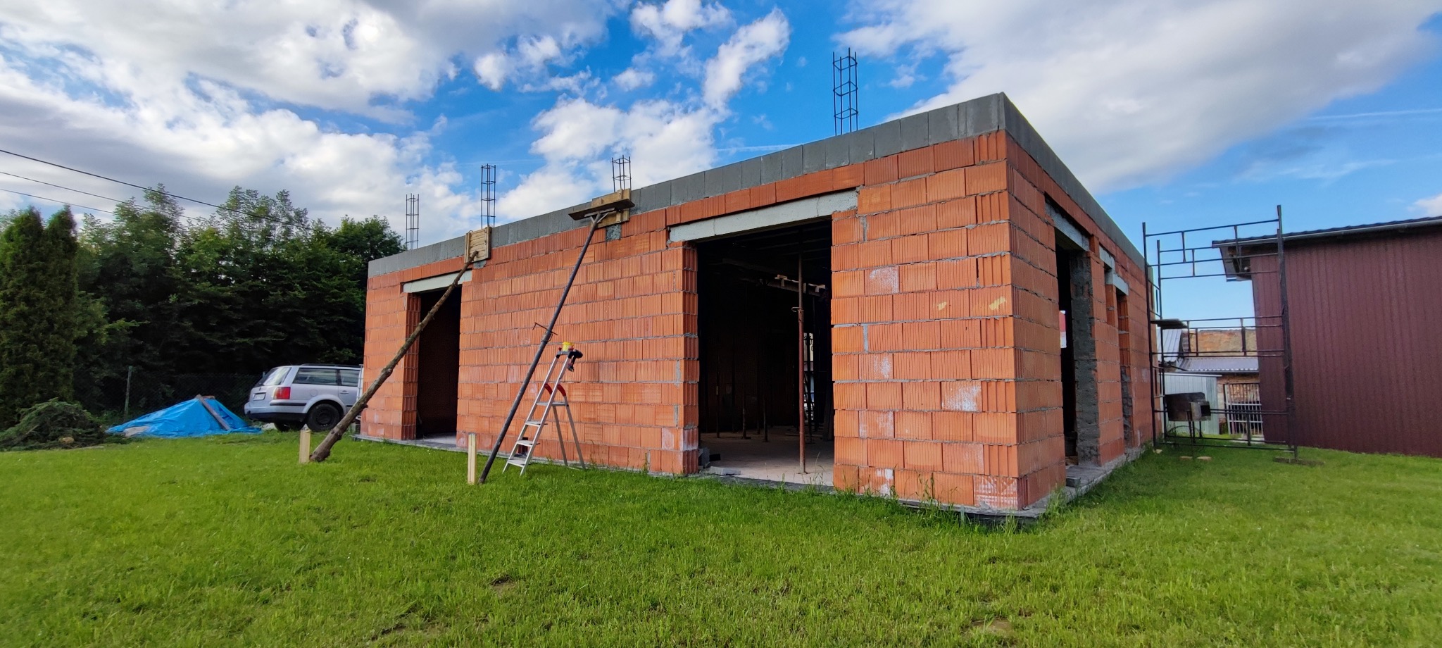 Budowa garażu z czerwonej cegły, widoczne zbrojenia na dachu, podparte ściany, drabina i rusztowanie. W tle zieleń i błękitne niebo.